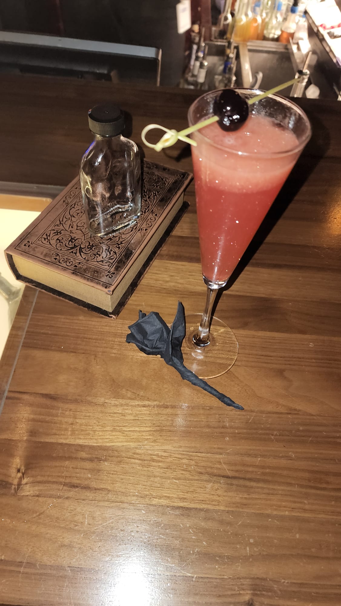 A pink cocktail garnished with a dark cherry on a skewer sits on a wooden bar next to an ornate book and an empty glass bottle. A black paper rose lies on the bar, adding a decorative touch.