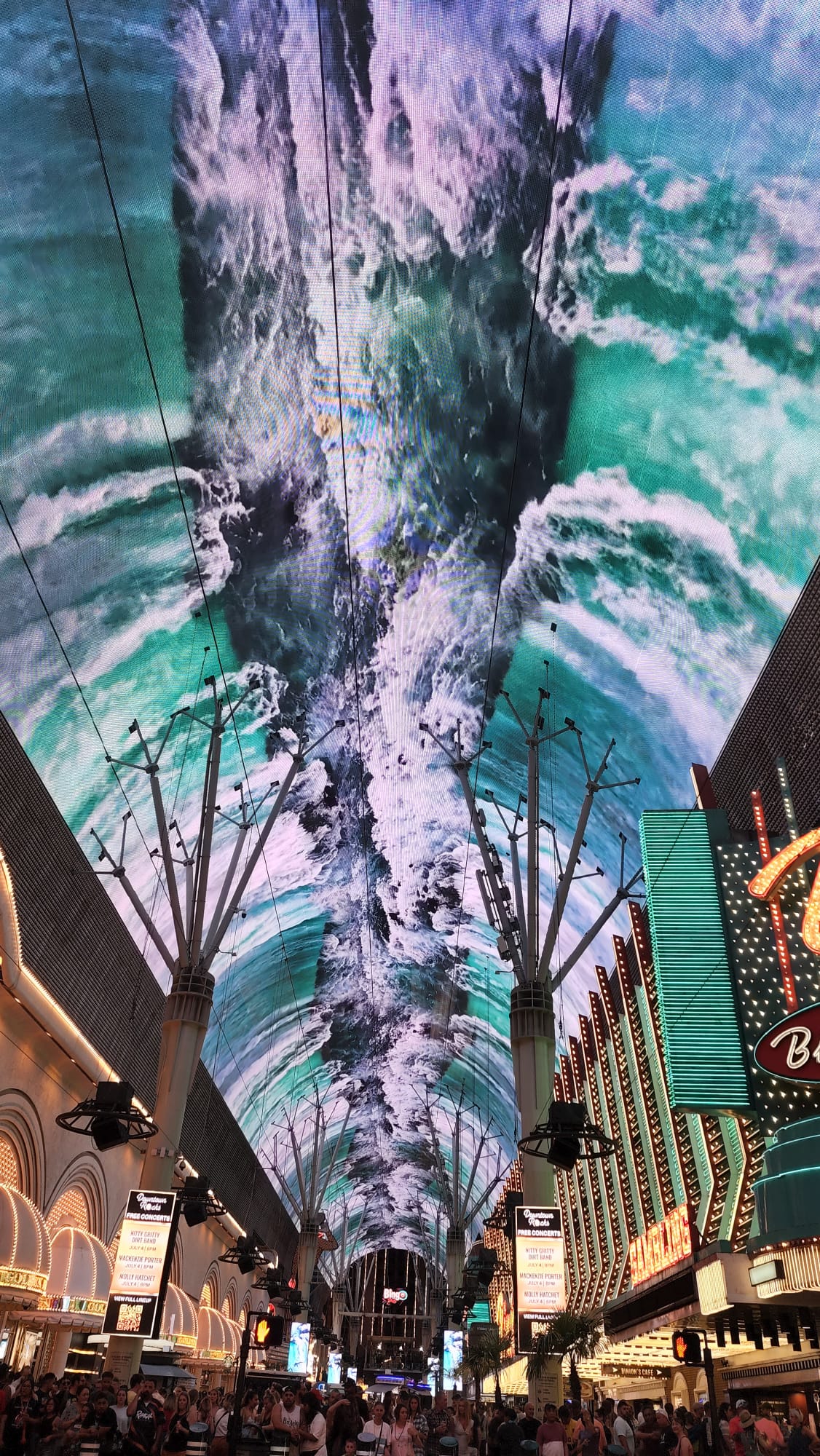 A bustling street scene under a massive, vibrant digital canopy displaying a dynamic ocean wave. The crowd below is lively, surrounded by illuminated signs and colorful neon lights on the buildings.