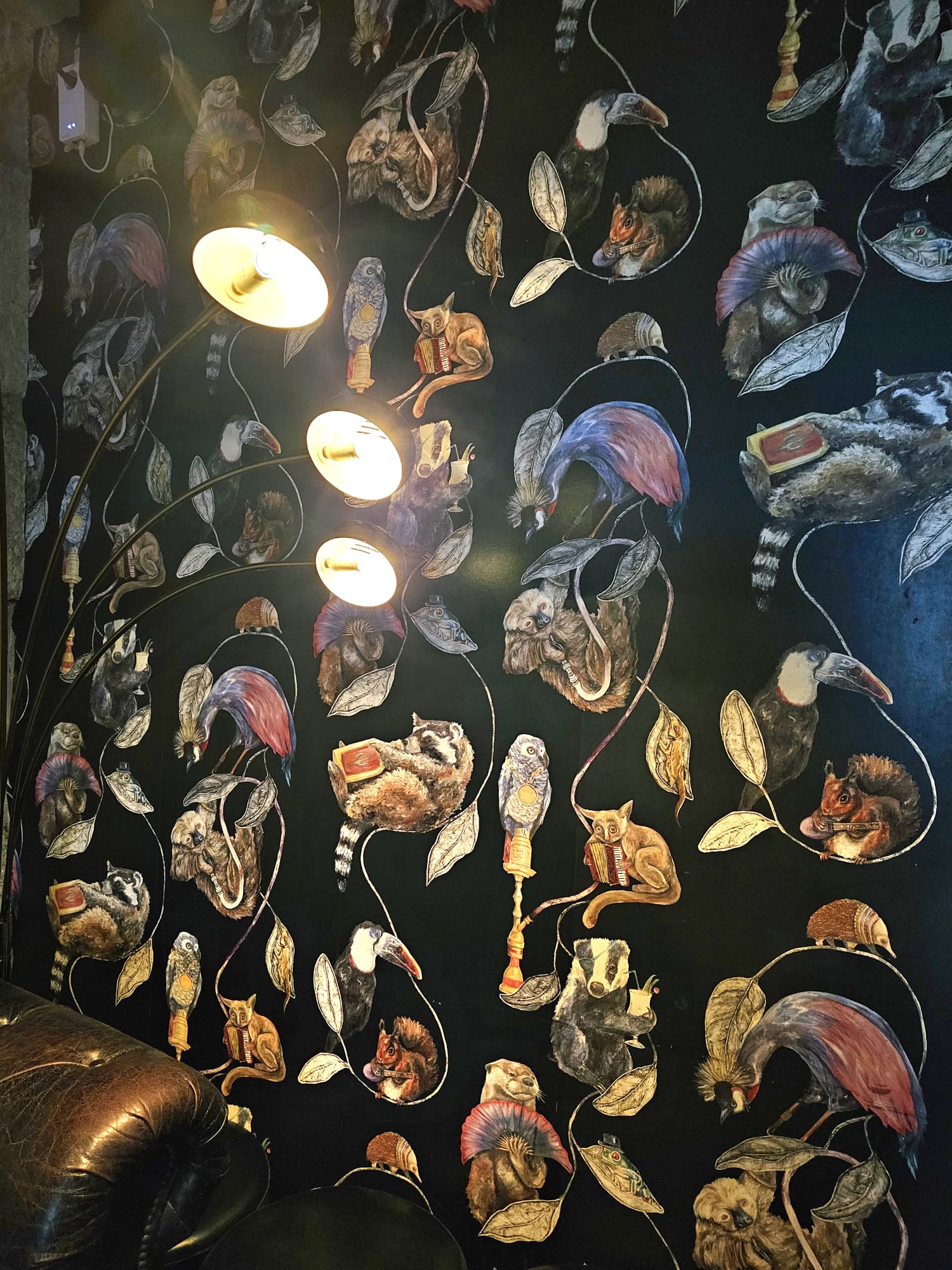 A whimsical wallpaper features various animals, such as owls, squirrels, and birds, against a dark background, each holding or interacting with objects like books and fans. A three-bulb floor lamp emits a warm glow, illuminating part of the scene, while a brown leather chair sits beneath.