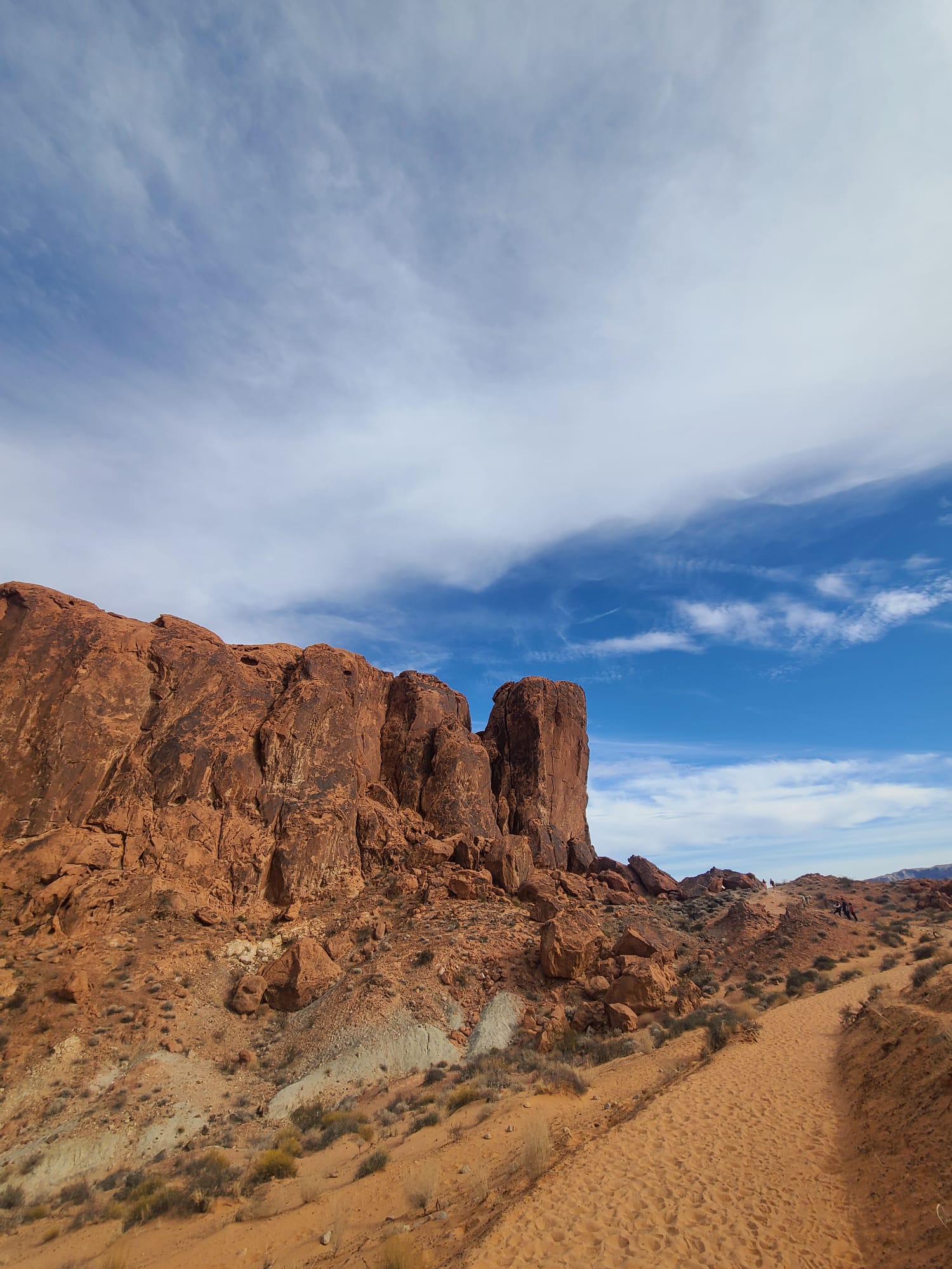 A rocky desert landscape features towering reddish-brown cliffs under a vast sky filled with wispy clouds. A sandy path winds through the arid terrain, dotted with sparse vegetation.