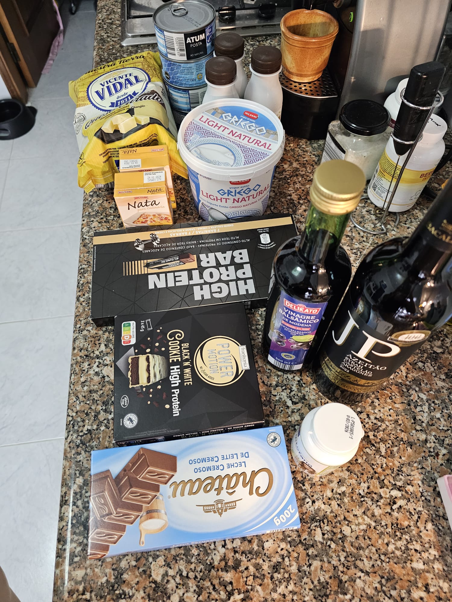 A variety of groceries are displayed on a kitchen counter, including canned tuna, bags of chips, a tub of Greek yogurt, bottles of vinegar and wine, a box of high-protein bars, chocolate, and small cartons labeled "Nata." The items are arranged neatly, showcasing diverse food products.