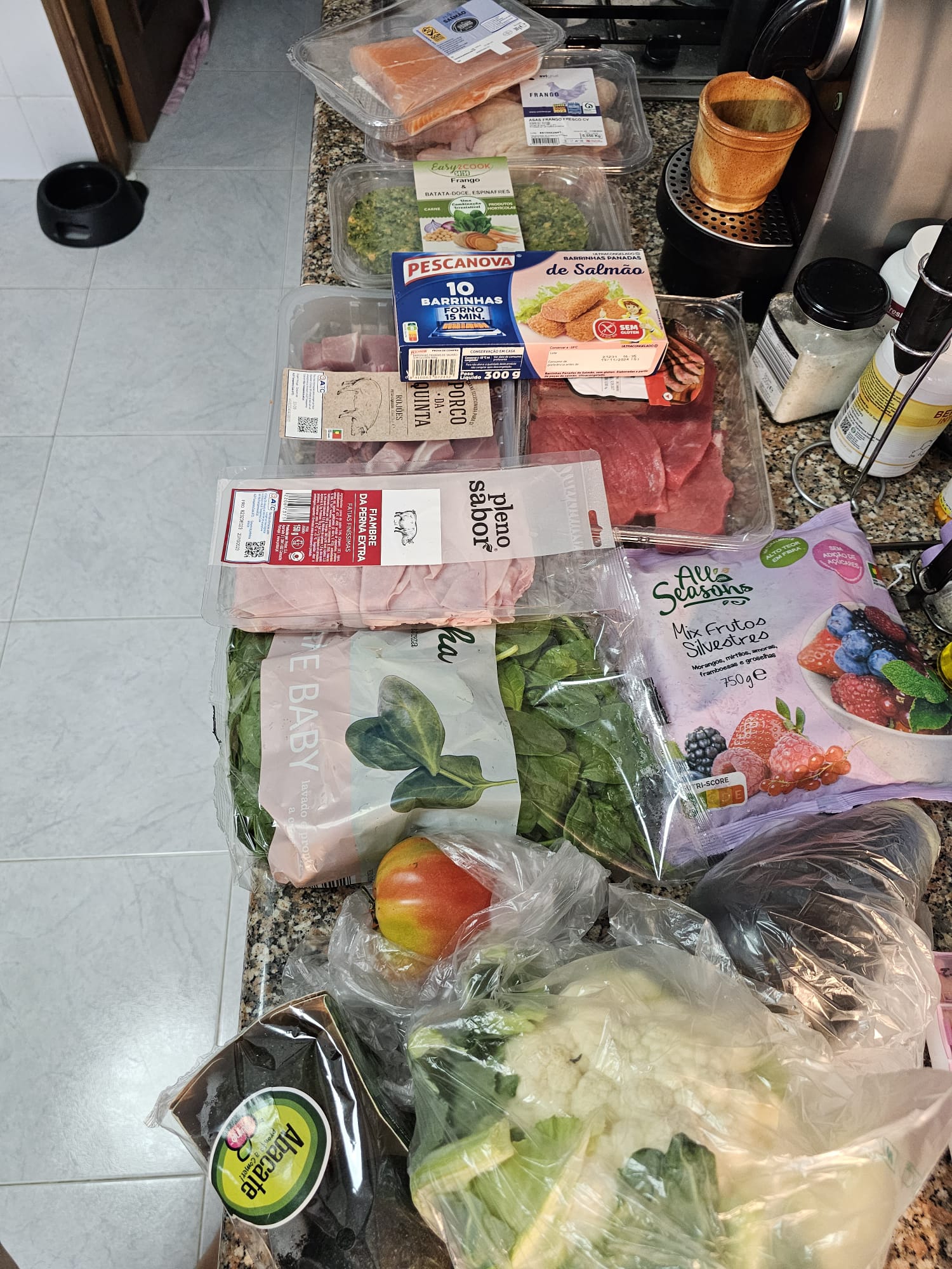 A kitchen counter filled with various groceries, including packaged salmon, pork, and beef, a box of breaded salmon bites, and frozen berries. Fresh produce such as spinach, a mango, cauliflower, and an avocado are also visible. Kitchen appliances and containers are in the background.