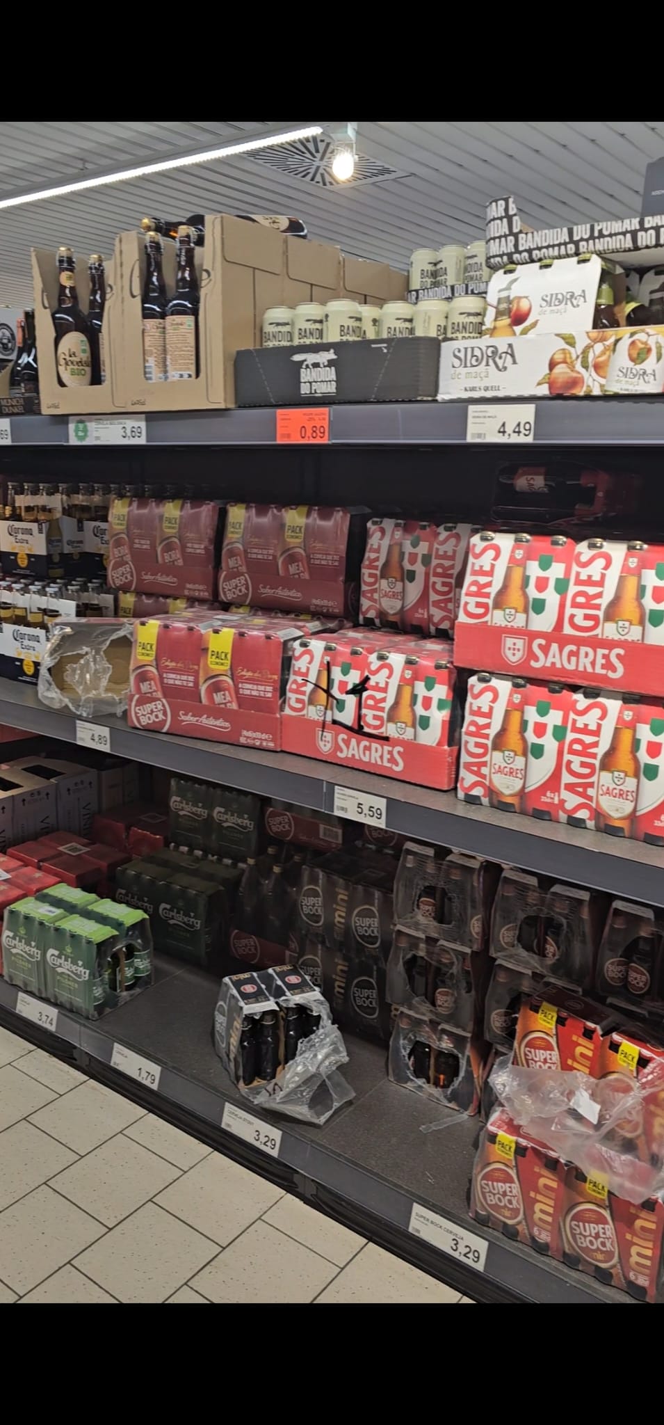 A supermarket aisle displays various beer and cider brands on shelves, including Sagres, Super Bock, and Carlsberg. The products are neatly arranged in packs with visible price tags, set against a tiled floor and under bright lighting.