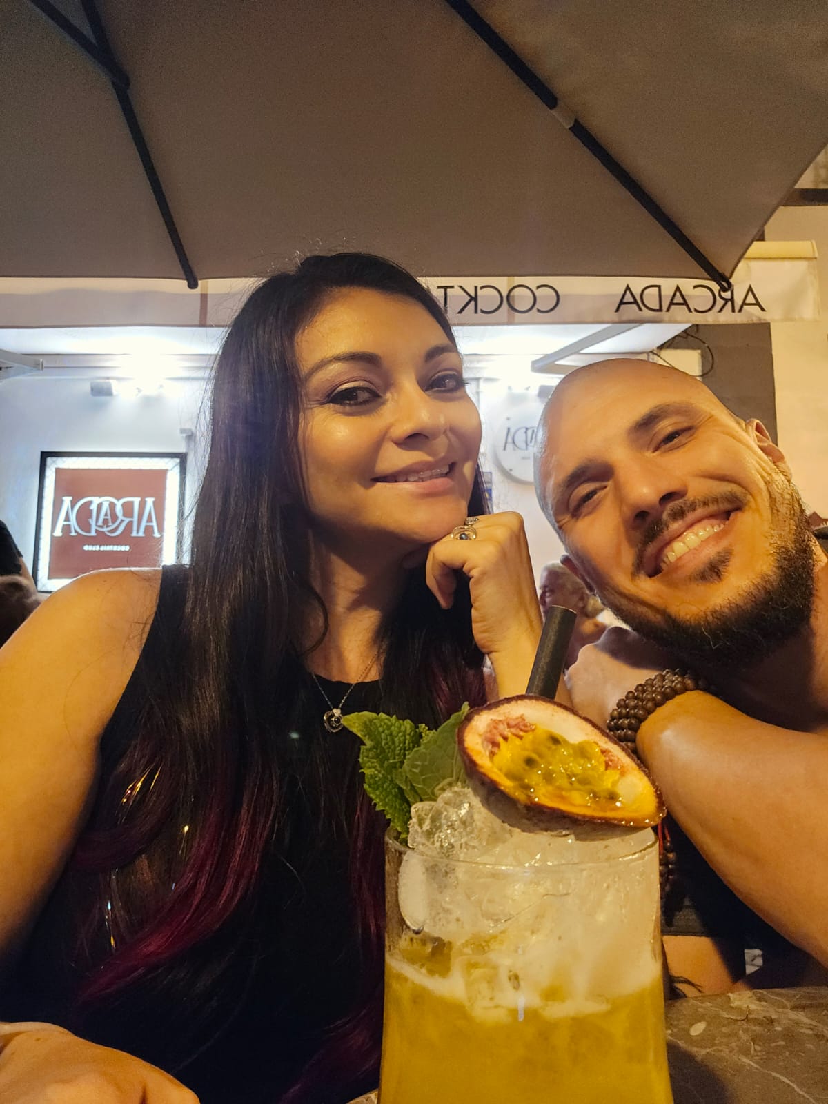 A smiling woman and man are seated at an outdoor café table, with a refreshing cocktail garnished with passion fruit and mint in the foreground. The café's sign is visible in the background under a large umbrella.