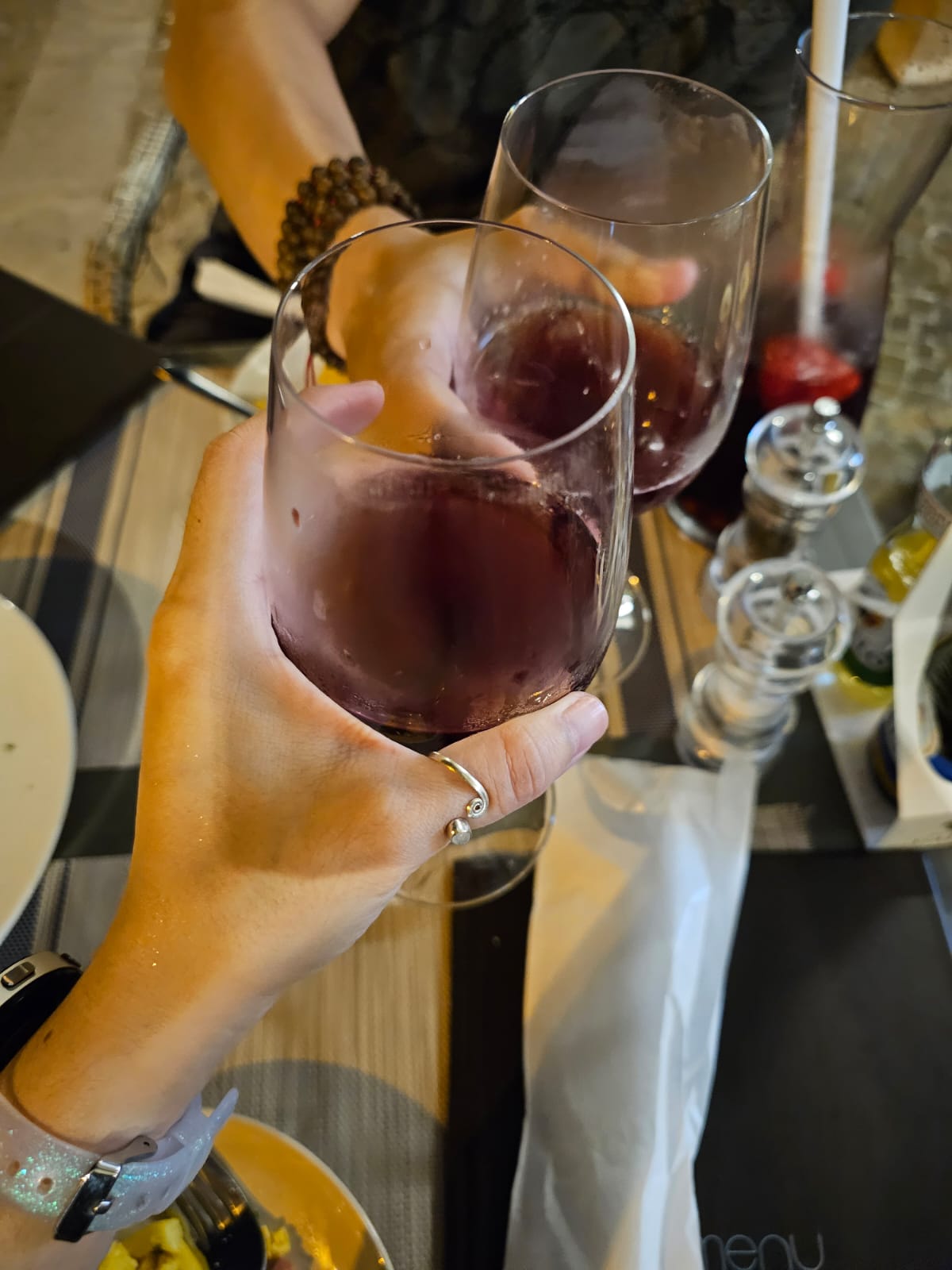 Two people clink glasses of red wine over a table set with a menu, pepper grinder, and condiments. One person wears a smartwatch and a ring, while the other has a beaded bracelet.