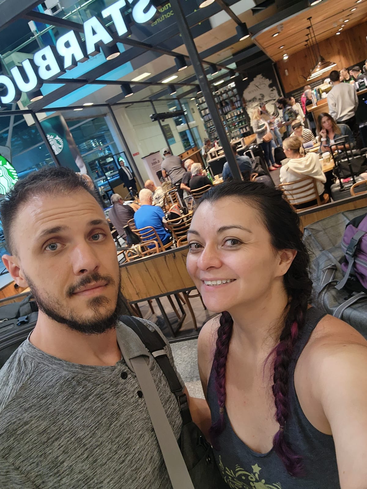 A man and woman take a selfie in front of a busy Starbucks, with the woman smiling and the man looking at the camera. Inside the cafe, people are seated at tables, enjoying their drinks in a lively atmosphere.