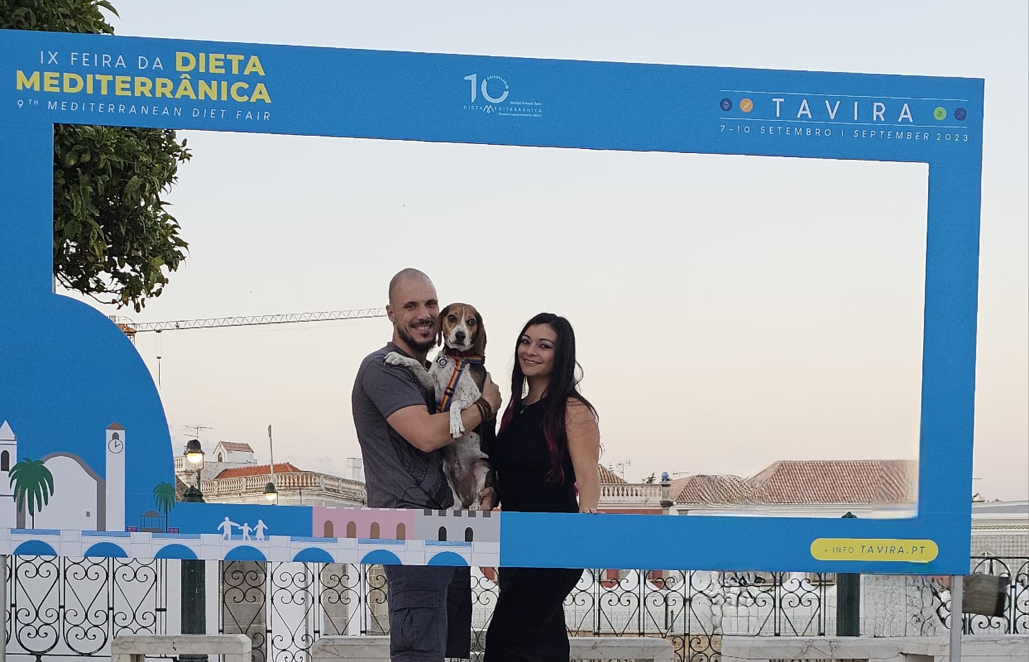 A man and woman smile while holding a dog, posing within a large blue photo frame promoting the "9th Mediterranean Diet Fair" in Tavira. The background shows a clear sky and rooftops, adding a scenic touch.