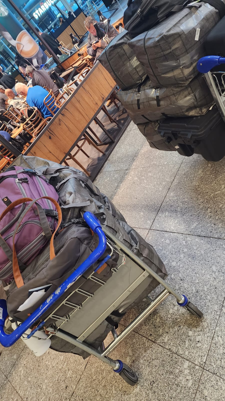 A luggage cart filled with several large, taped suitcases and a purple backpack is in an airport setting, with a Starbucks visible in the background and people seated at nearby tables.