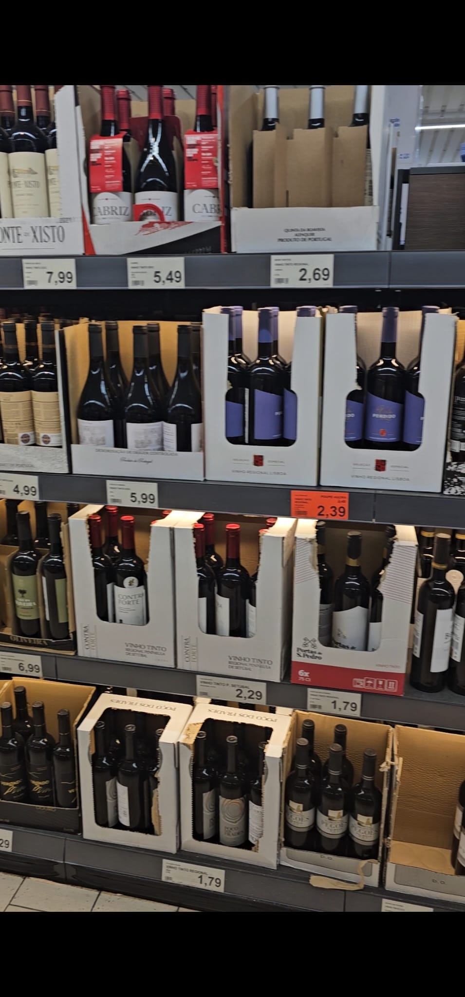 A supermarket wine aisle displays various bottles of red wine in cardboard boxes, with visible price tags ranging from 1.79 to 7.99. The shelves are stocked with different brands, featuring labels in various colors, predominantly red and purple.