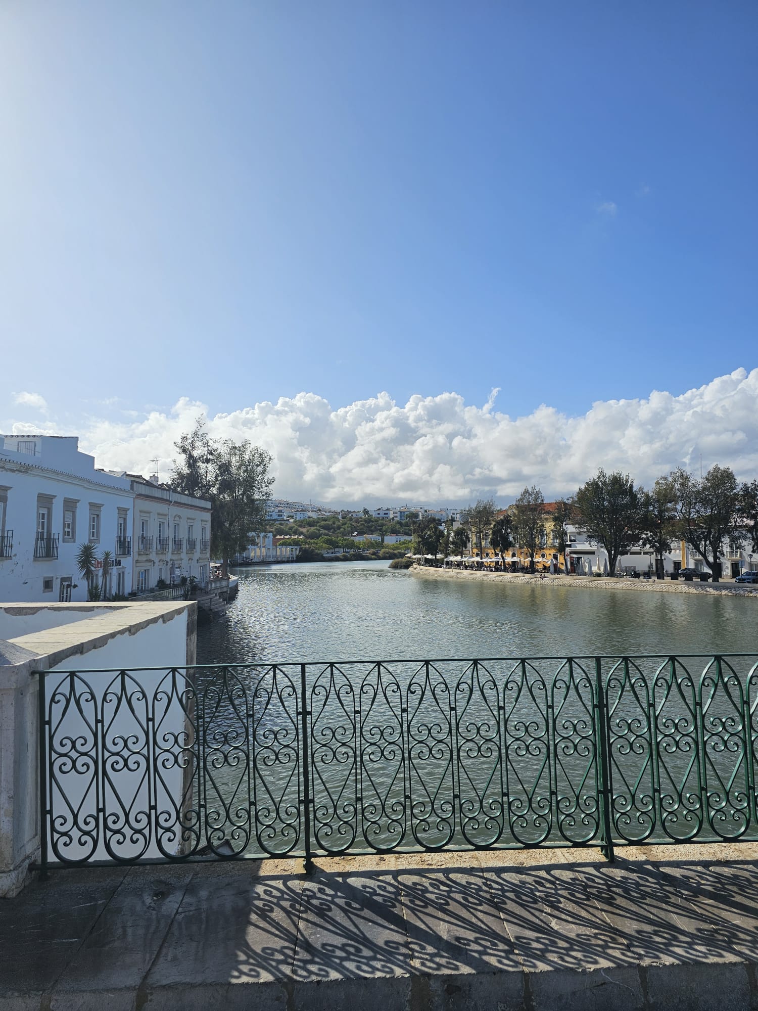 A scenic view of a river bordered by elegant wrought iron railings, with white buildings lining the left bank and trees on the right. The sky is clear and blue, dotted with fluffy white clouds, creating a serene atmosphere.