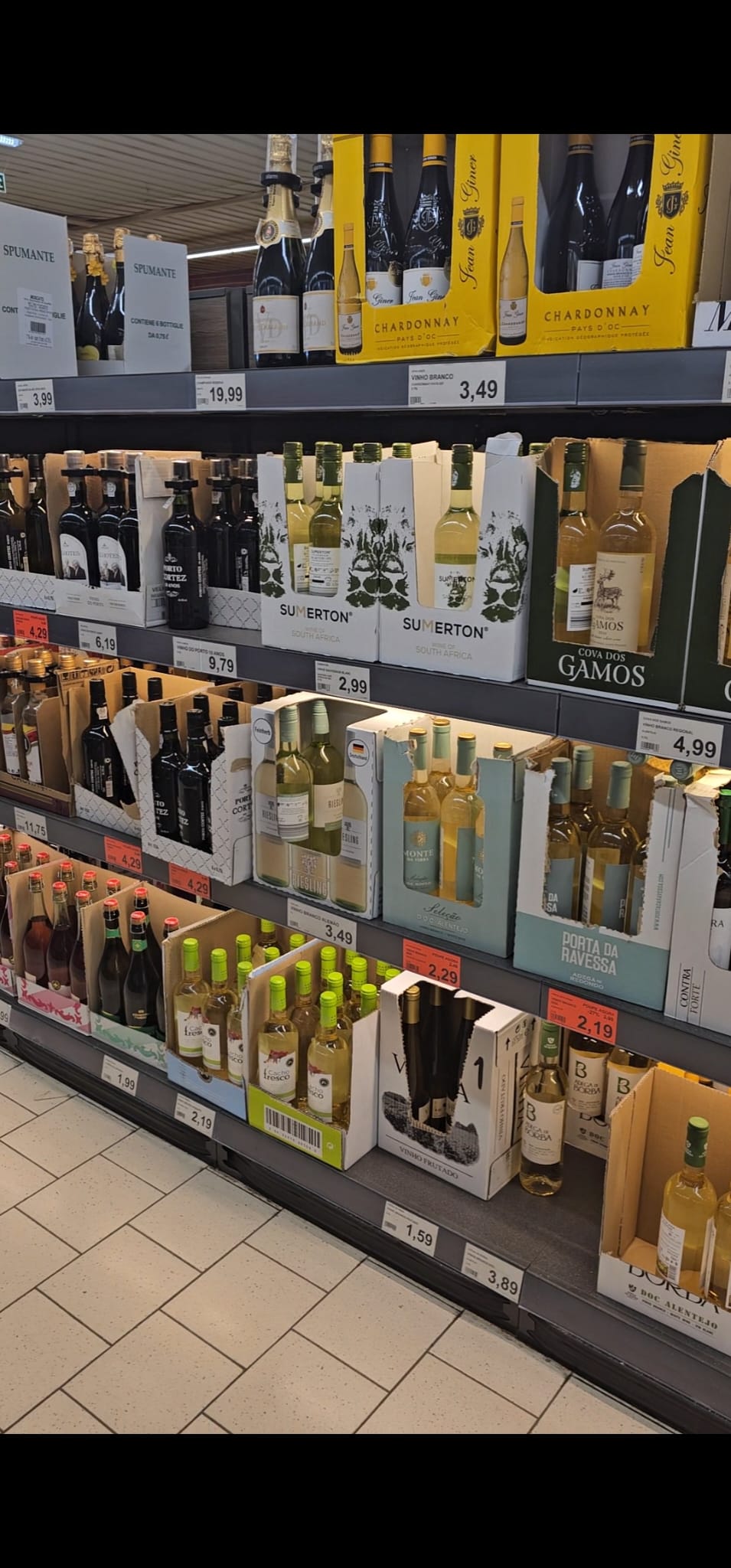 A supermarket shelf displays a variety of wines, including bottles of sparkling wine, Chardonnay, and other white wines. The bottles are organized in cardboard boxes with visible price tags, ranging from €1.59 to €19.99. The setting is well-lit, highlighting the different labels and colors of the wine bottles.