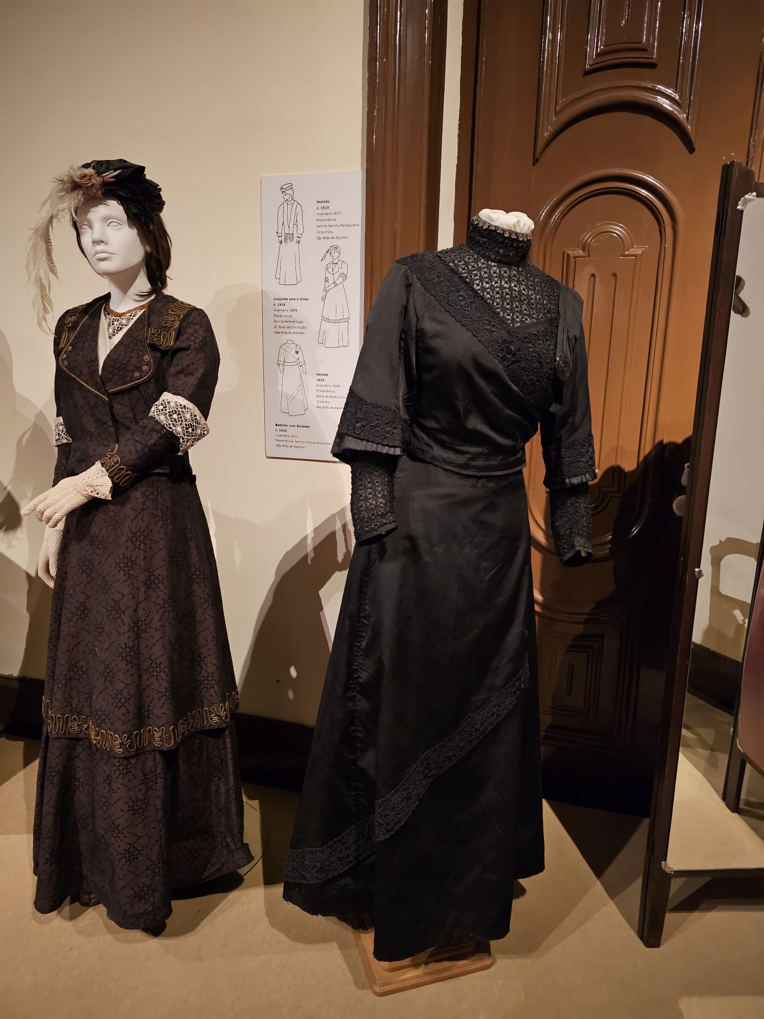 Two mannequins display elegant historical dresses in a museum setting. The left mannequin wears a dark patterned dress with lace cuffs and a decorative hat featuring feathers. The right mannequin showcases a black dress with intricate lace detailing and a high collar. A poster with dress illustrations is visible on the wall behind them.