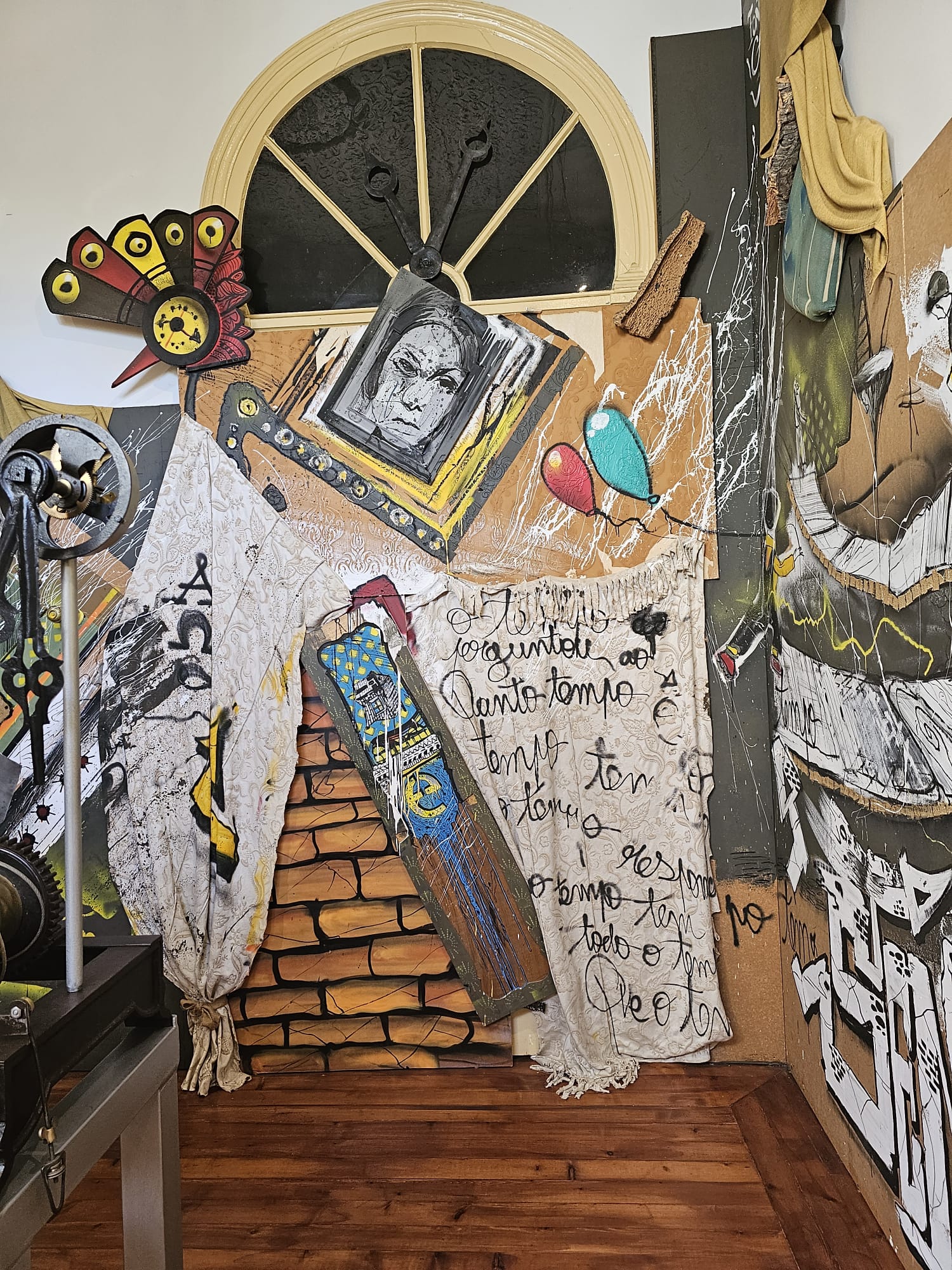 A vibrant art installation features a mix of painted and three-dimensional elements, including a large abstract face, colorful balloons, and a clock-like design. The background resembles a brick wall, partially covered by draped fabric with handwritten text. The setting includes a wooden floor and a semicircular window at the top.