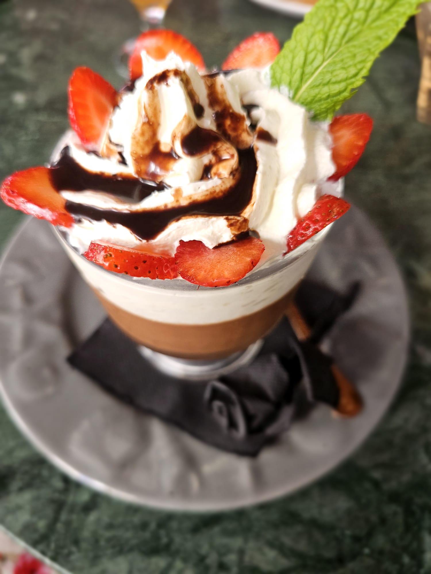A glass of chocolate panna cotta with whipped cream, strawberries, chocolate sauce and a mint leaf