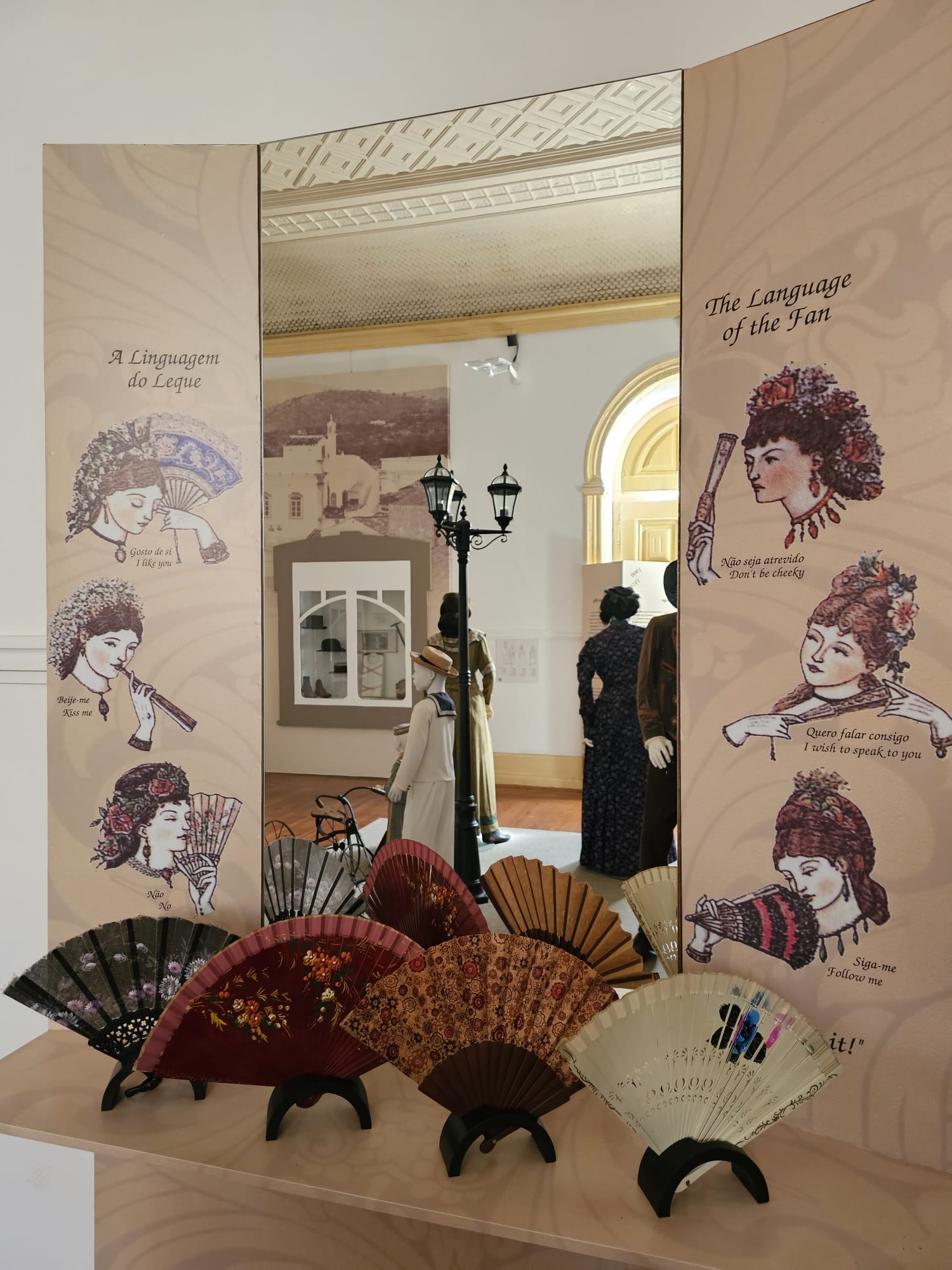 A display showcases five decorative hand fans with intricate patterns and vibrant colors. Behind them, a panel illustrates "The Language of the Fan," depicting vintage-style illustrations of women using fans, each with a caption explaining a gesture's meaning. The setting includes mannequins dressed in historical attire, adding to the vintage ambiance.