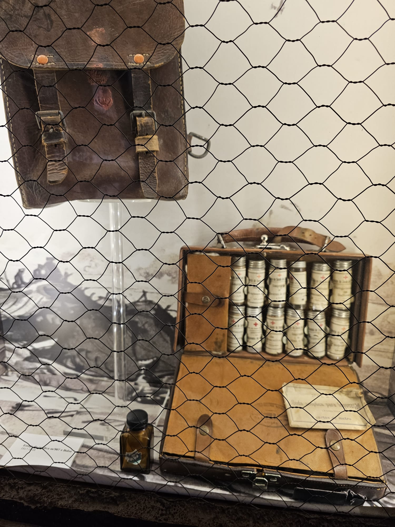 A vintage display shows a worn leather satchel and an open case filled with neatly arranged glass vials, each with a label and red cross. A small brown bottle and a piece of paper lie beside the case, all behind a wire mesh.