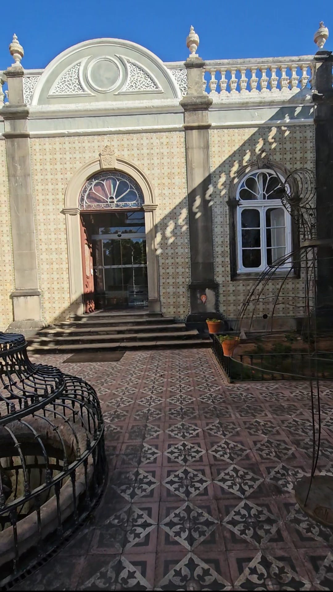 A historic building facade with ornate tile patterns and arched windows, featuring a decorative wrought iron structure in the foreground. The entrance has a set of steps leading up to a doorway with a semicircular stained glass window above. The tiled courtyard displays intricate geometric designs under a clear blue sky.