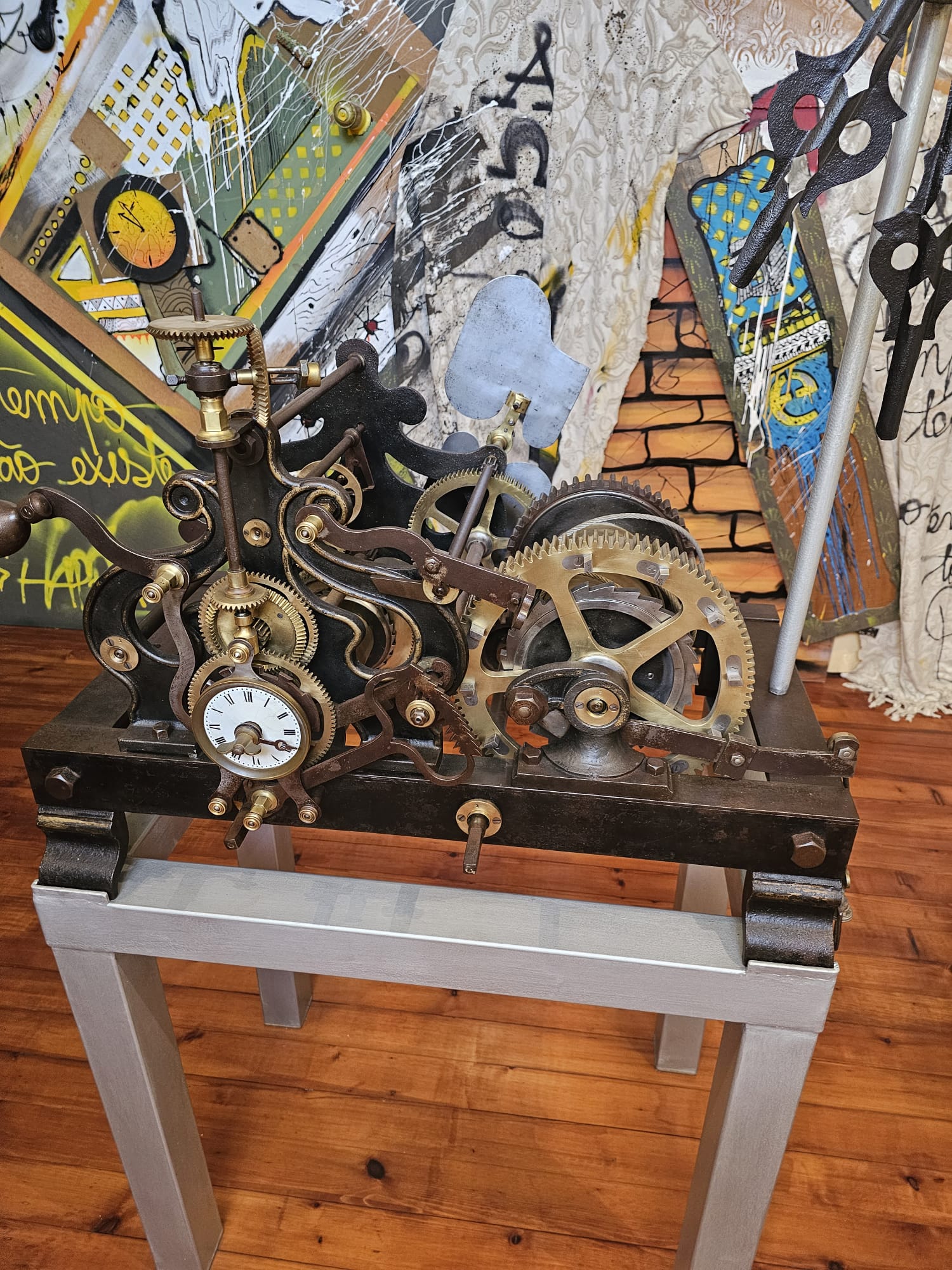 An intricate antique clock mechanism with visible gears and brass components is displayed on a sturdy metal stand. Behind it, a colorful, graffiti-style mural adds a vibrant backdrop to the scene.