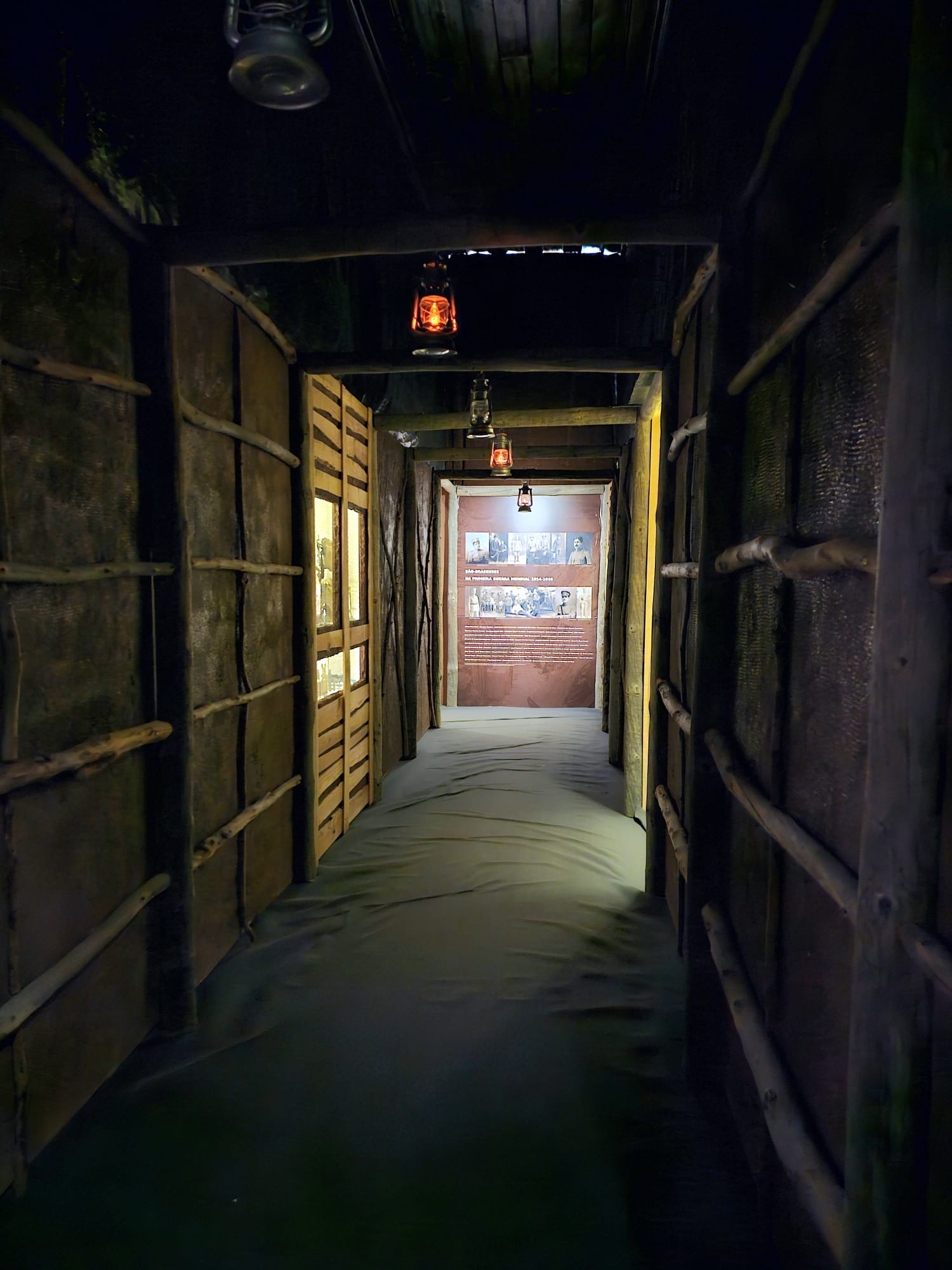 A dimly lit corridor with wooden walls and beams, featuring hanging lanterns casting a warm glow. At the end, a display board with historical photos and text is visible, enhancing the vintage ambiance.