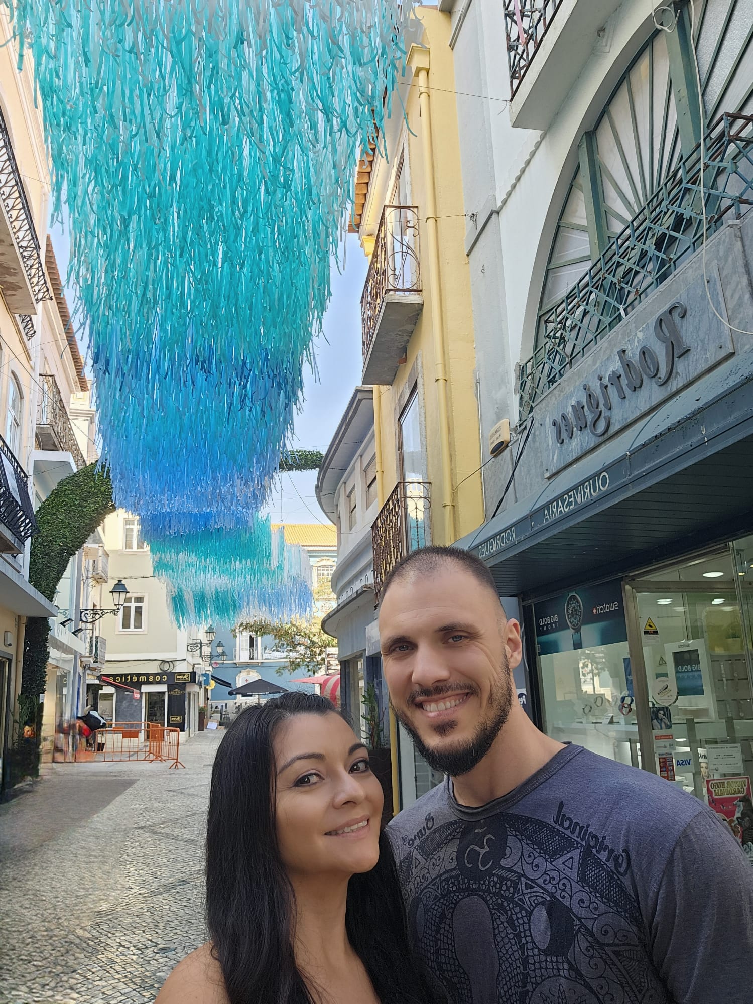 a couple smiling in the middle of a cobblestone, Portuguese street with blue decorations hanging overhead
