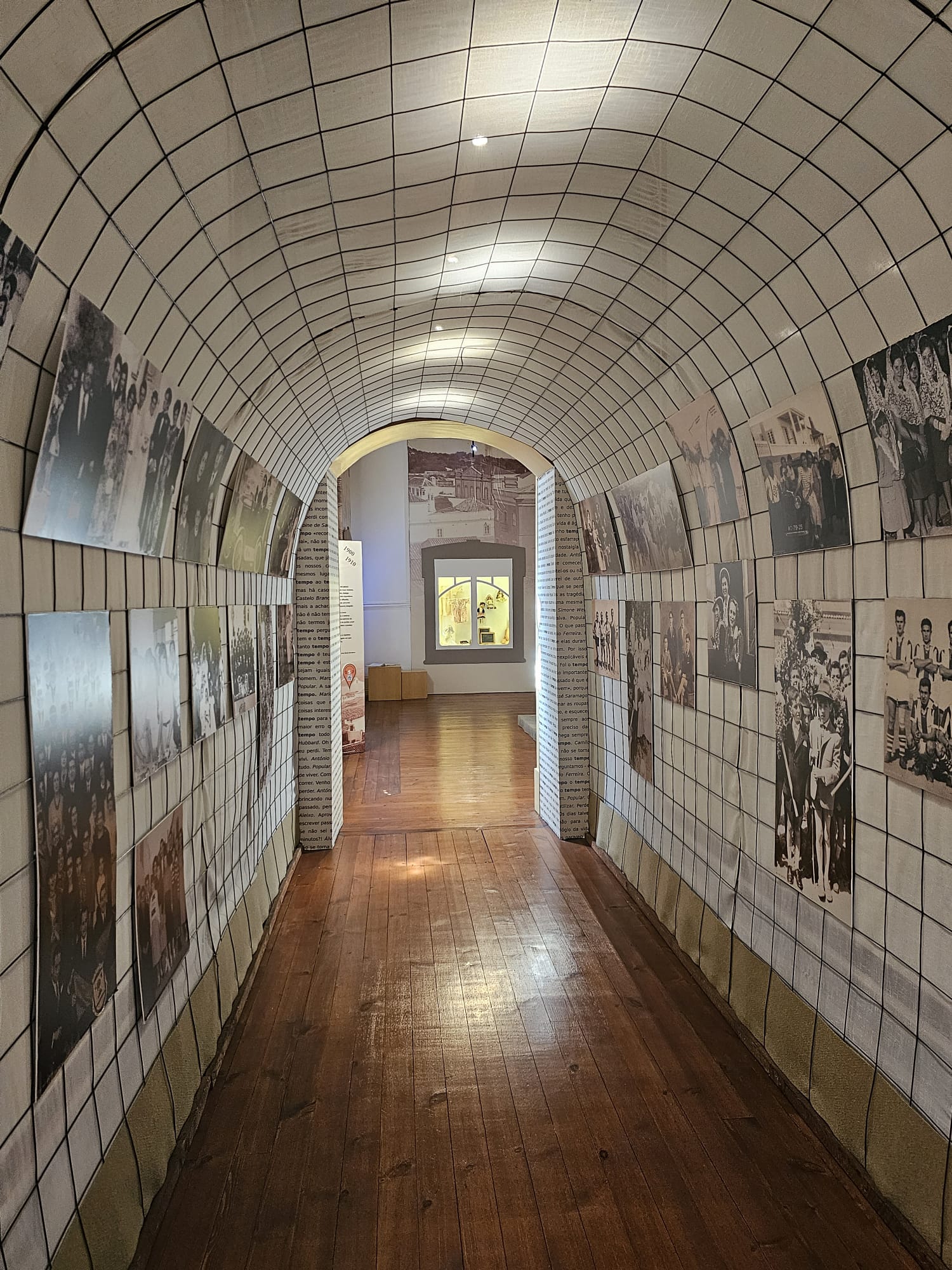 A corridor with a curved, grid-patterned ceiling and walls adorned with black-and-white photographs on both sides. The floor is wooden, leading to a well-lit room at the end, creating a historical and inviting atmosphere.