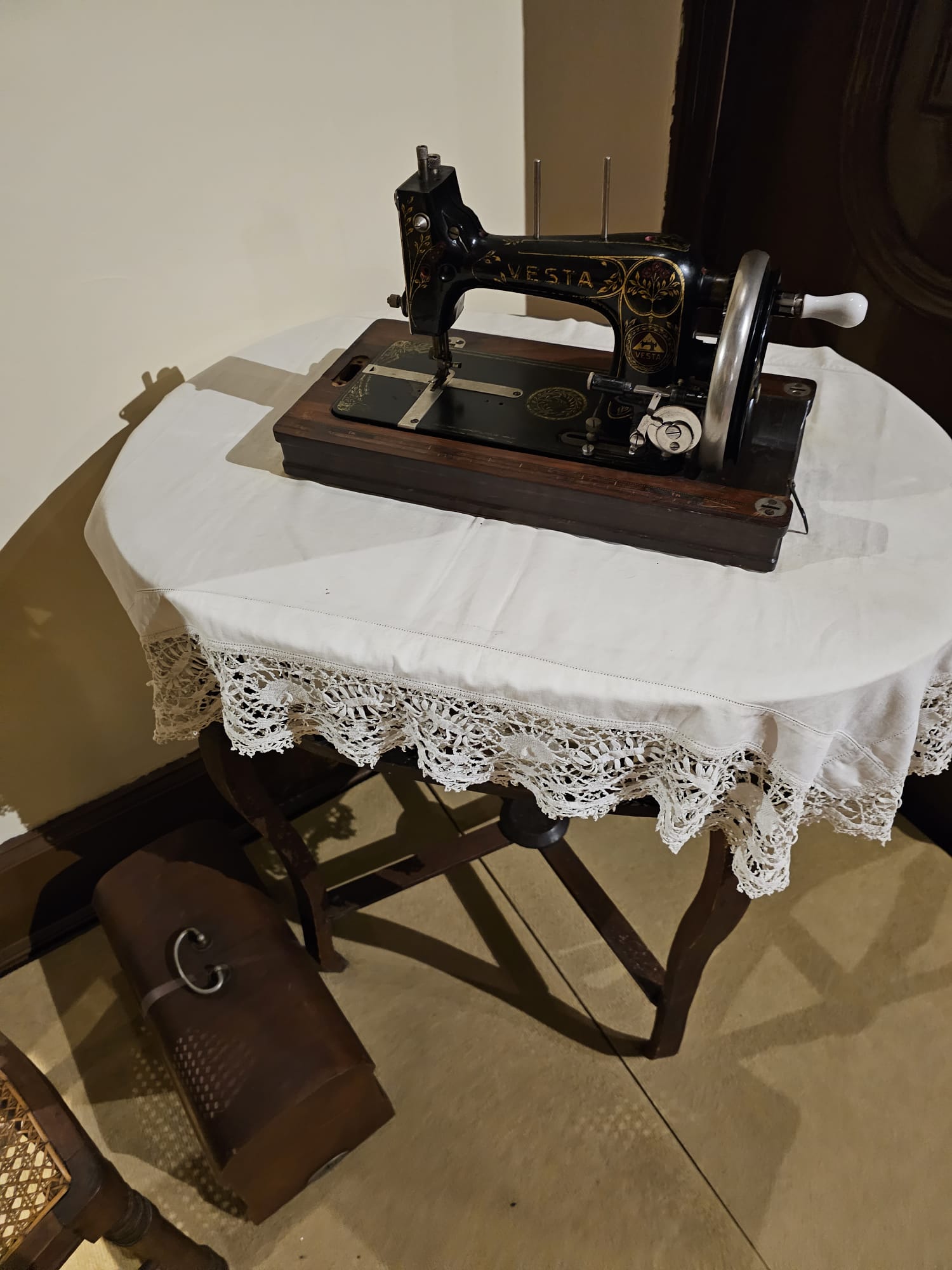 A vintage Vesta sewing machine sits on a small table covered with a lace-edged white cloth, with a wooden case resting on the floor nearby.