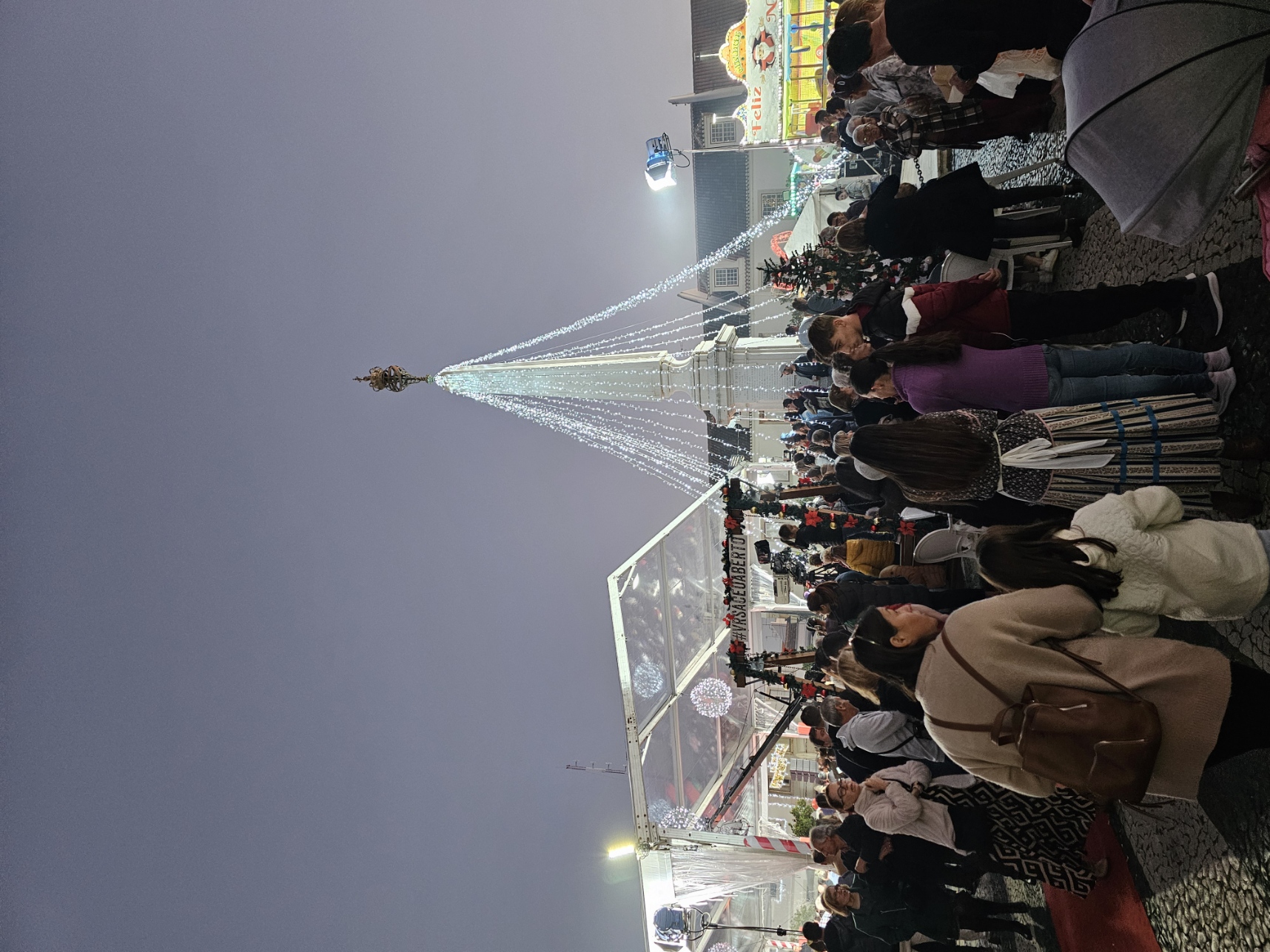 Large crowd gathered around the illuminated central light tree at the VRSA Christmas Village festival in Portugal at night