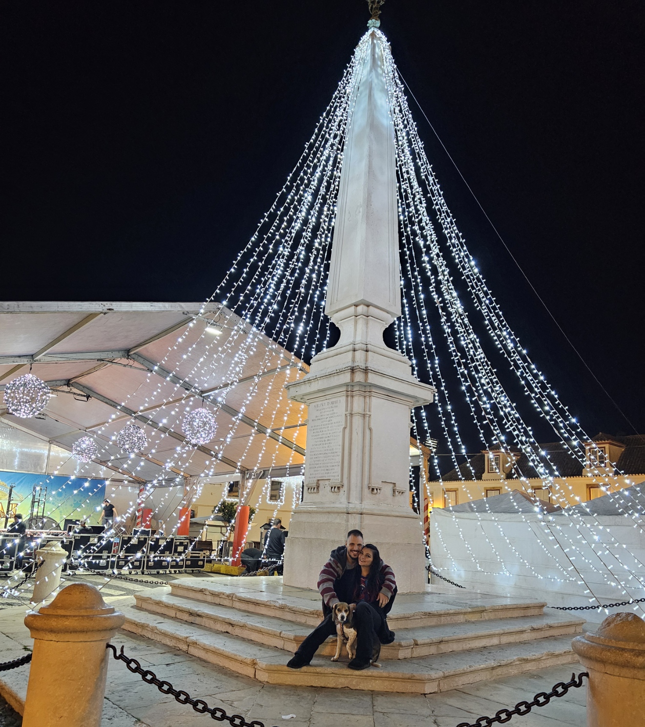 Couple sitting with their dog in front of a giant illuminated light tree at the VRSA Christmas Village in Vila Real de Santo António Portugal