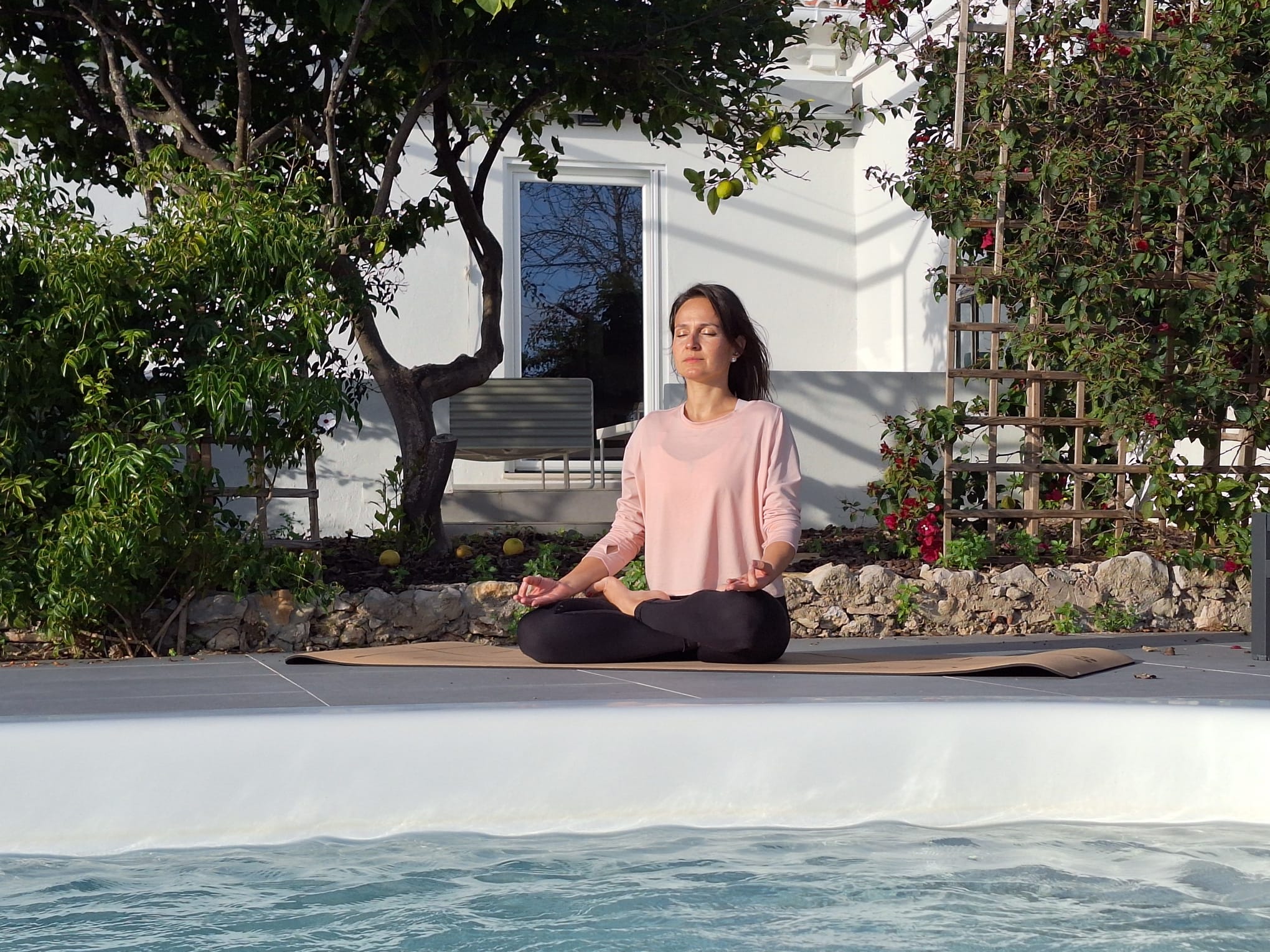 Woman sitting in a meditation pose on a yoga mat in the serene outdoor garden area of Human Movement Studio in Portugal