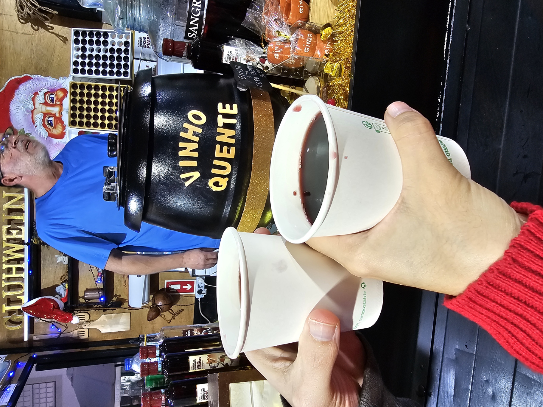 Two cups of vinho quente Portuguese mulled wine held up in front of a Gluhwein market stall at the VRSA Christmas Village