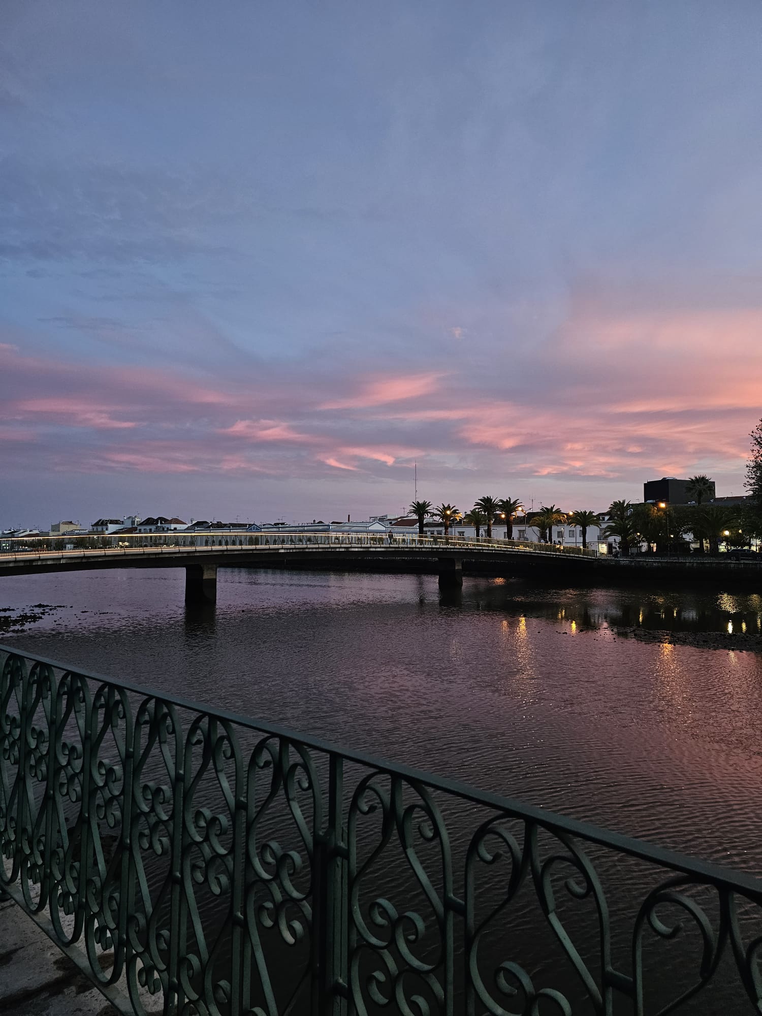 Pink and purple sunset over the Gilão River and bridge in Tavira, Portugal, with ornate iron railings in the foreground