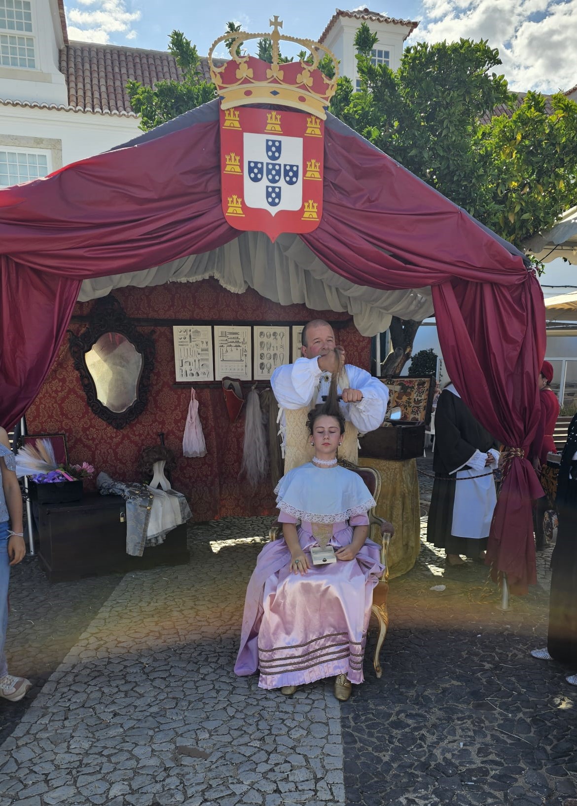Period costume hairdresser demonstration under a tent decorated with the Portuguese coat of arms at a historical festival