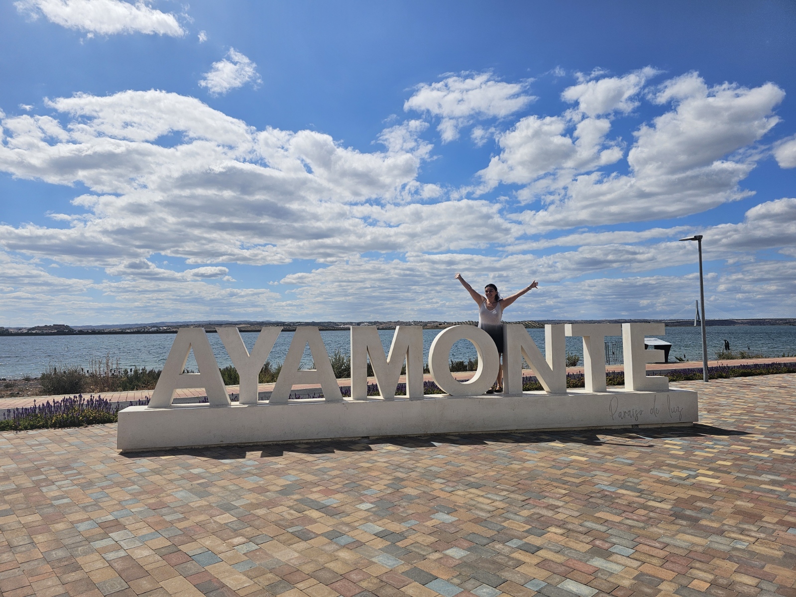 Woman standing on the large Ayamonte letter sign with the Guadiana River and blue sky in the background, Ayamonte Spain