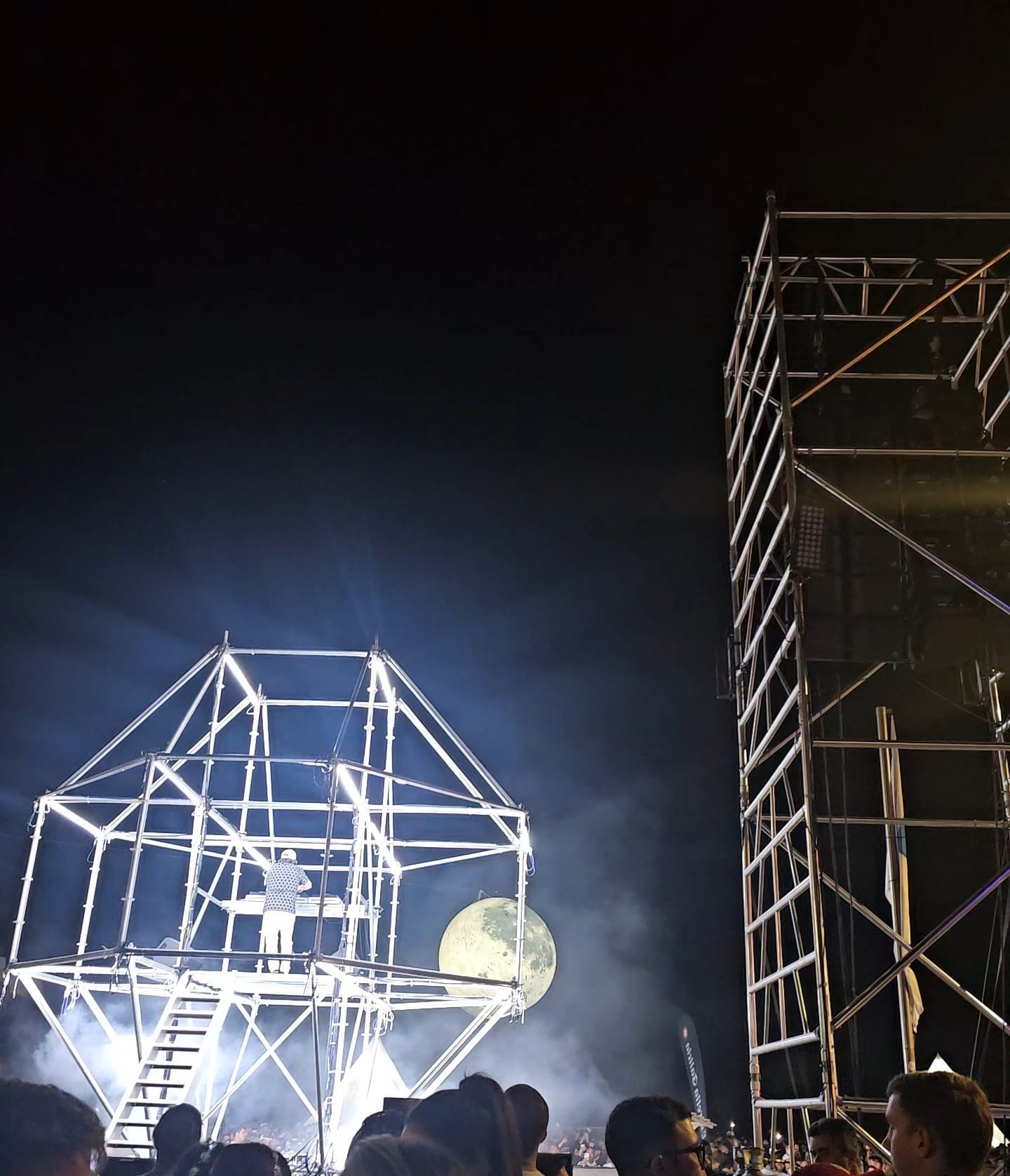 DJ performing on a geometric scaffold stage with a giant illuminated moon hanging over the crowd at Carvoeiro Black and White Night