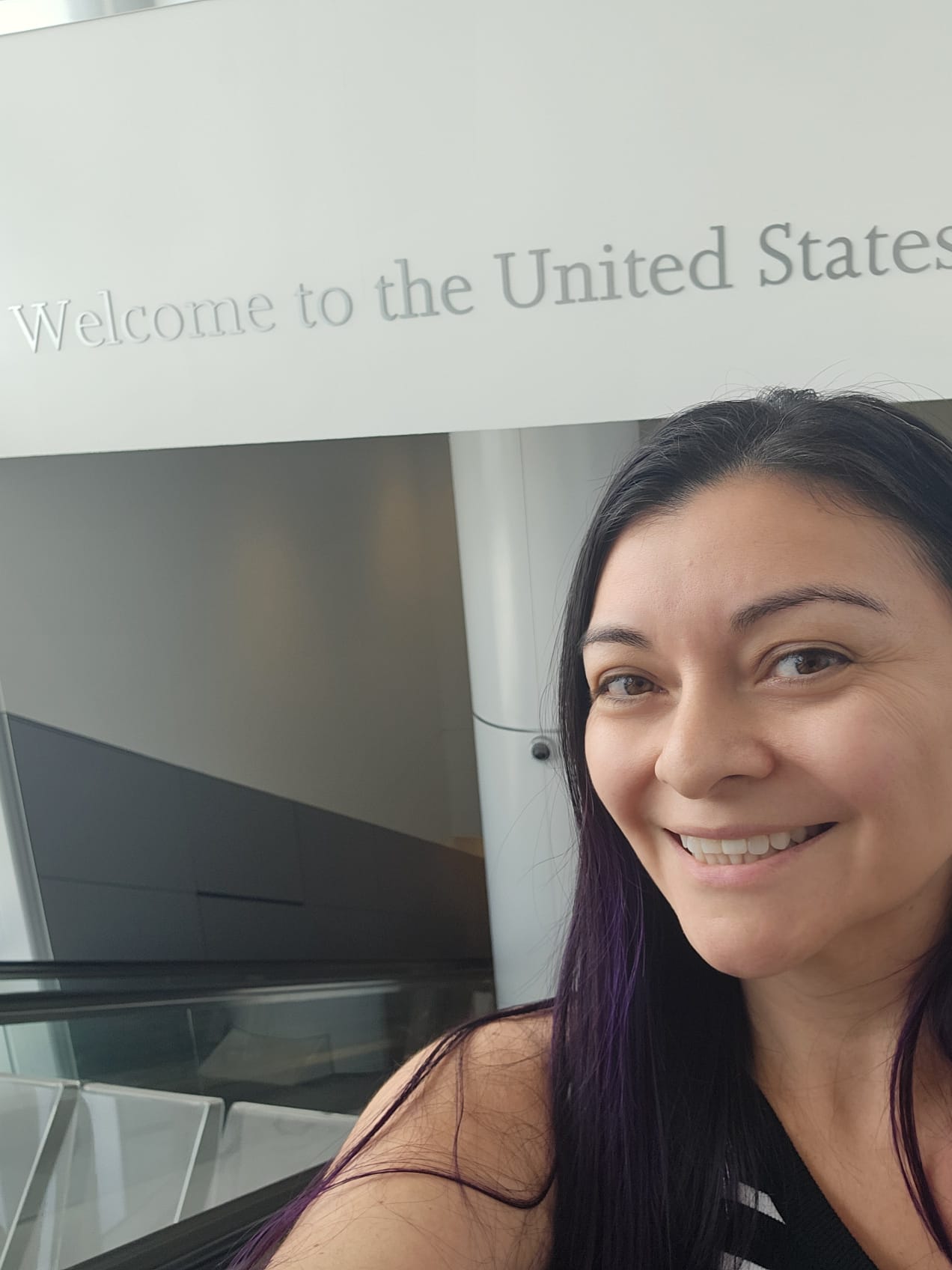 Woman with purple hair smiling in front of a Welcome to the United States sign at LAX airport after returning from Portugal