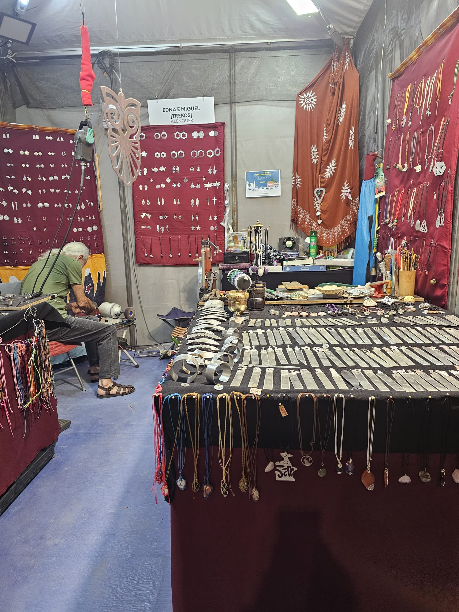 Artisan silversmith working at a craft booth displaying handmade silver jewelry, necklaces, and decorative pieces at an outdoor fair in Portugal