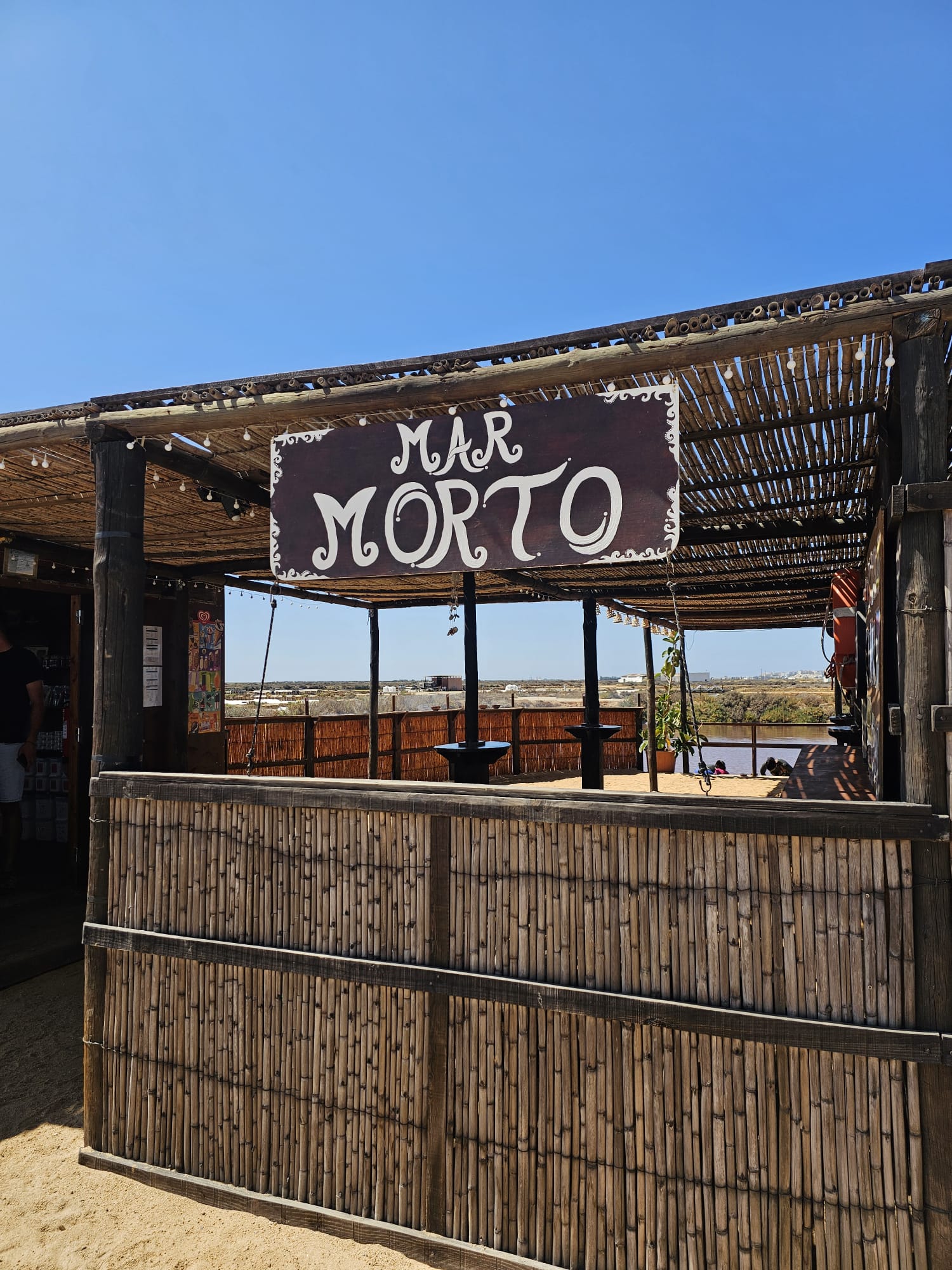 Rustic bamboo entrance sign reading Mar Morto at a salt flat spa in Olhao, Algarve, Portugal, under a bright blue sky