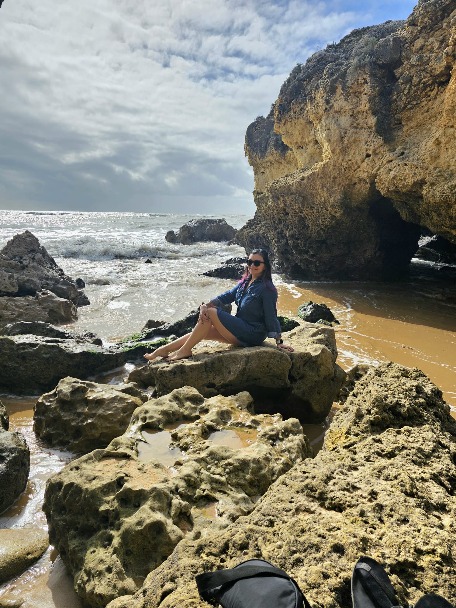 Woman with purple hair sitting on rocky coastal cliffs at Albufeira beach in Portugal with dramatic Atlantic Ocean waves and golden sandstone rocks in the background