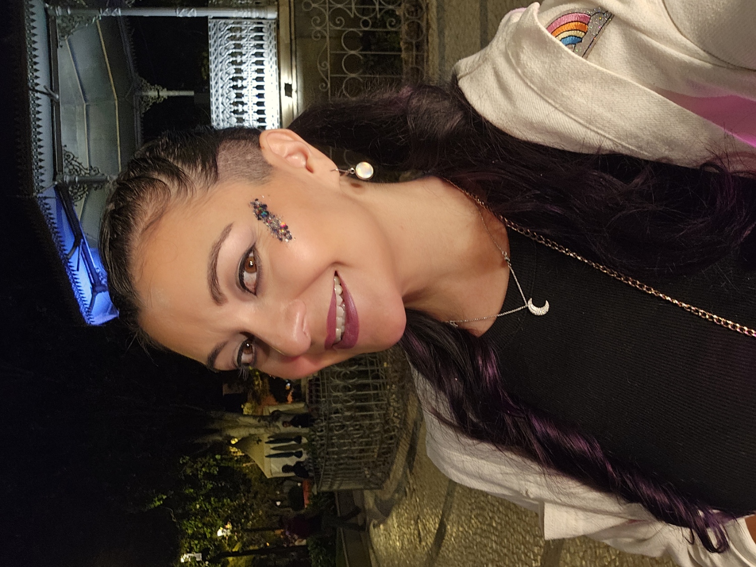 Woman with star-shaped face glitter and purple hair smiling at night in Tavira's town square during Festa da Juventude festival in Portugal