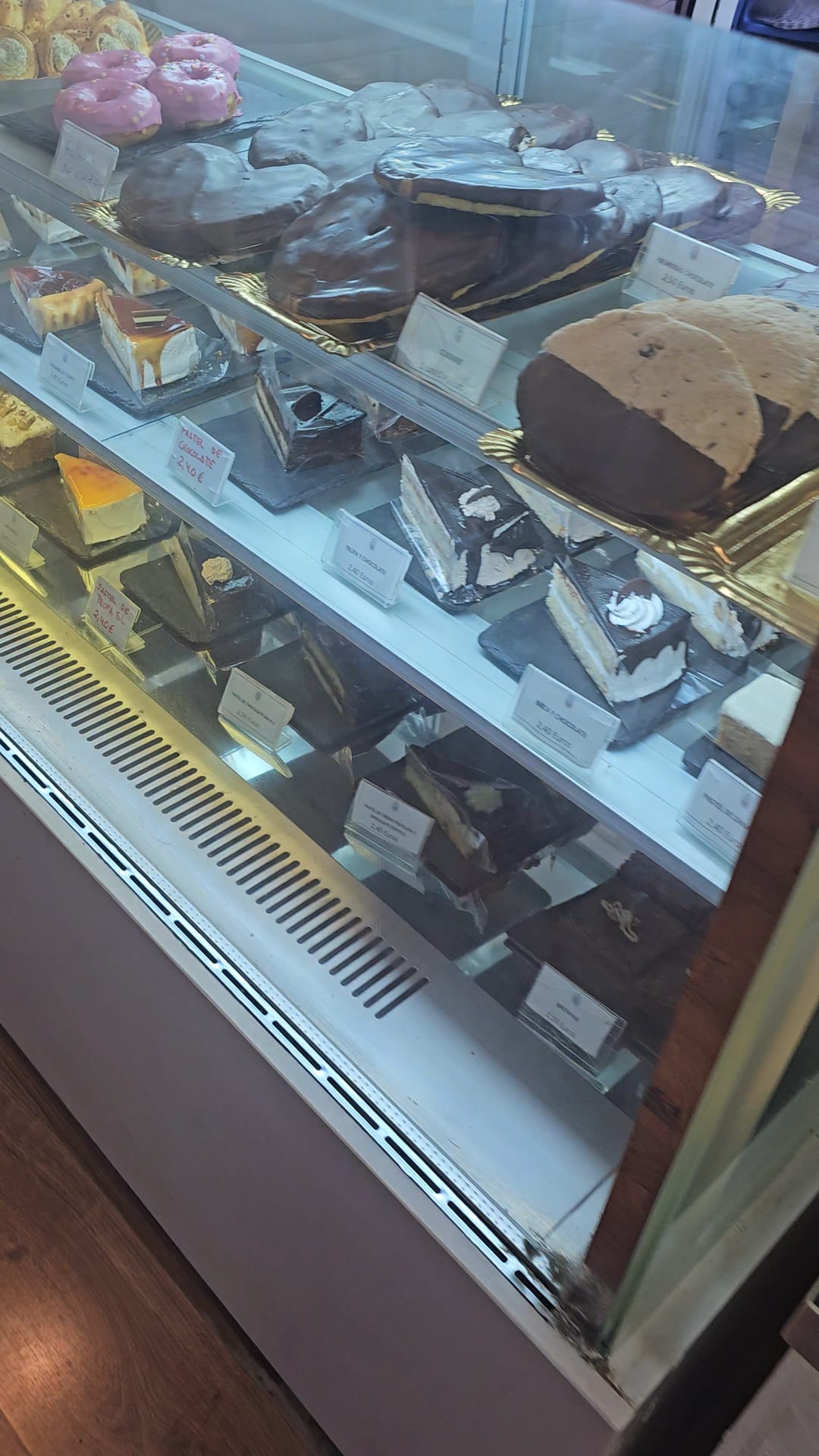 A large glass display case filled with a variety of desserts. The top shelf holds pink frosted donuts and large chocolate-dipped pastries, while the lower shelves feature assorted cake slices, including cheesecake, chocolate cake, and cream-filled layers, each with individual price tags.