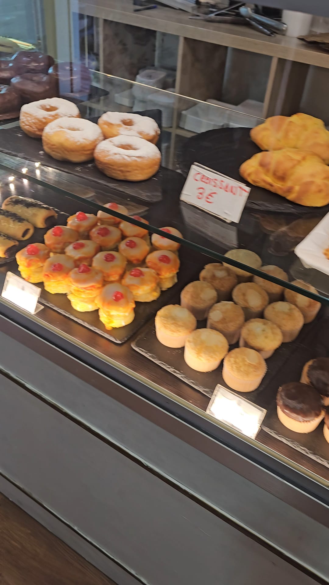 A wooden counter displaying several trays of gluten-free pastries, including sugar-dusted donuts, golden croissants, small muffins, and bite-sized cakes with red toppings. A small hand-written sign indicates croissants for €3.