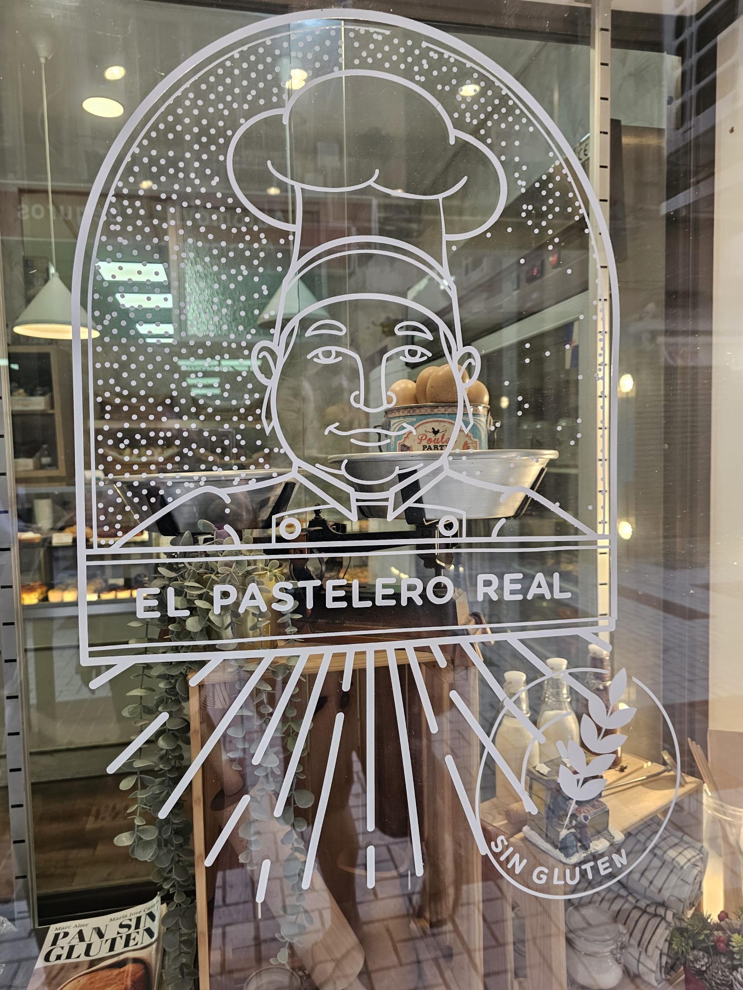 glass door or window featuring the white outline logo of "El Pastelero Real," which includes a caricature of a chef wearing a toque. The logo is positioned above a "Sin Gluten" (gluten-free) wheat symbol.