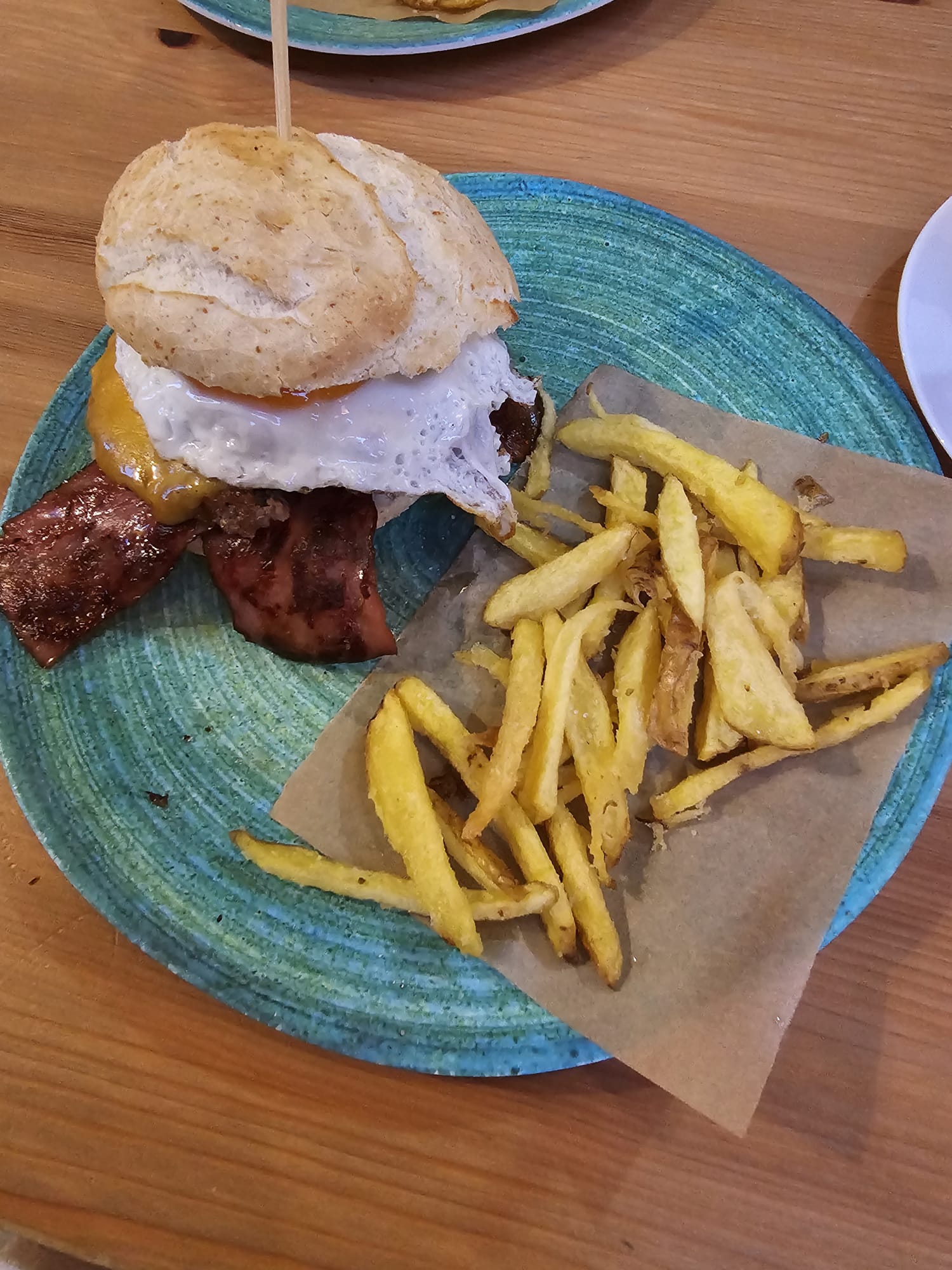 A high-angle shot of a massive gluten-free burger with a fried egg and bacon, served with a side of thick-cut french fries.