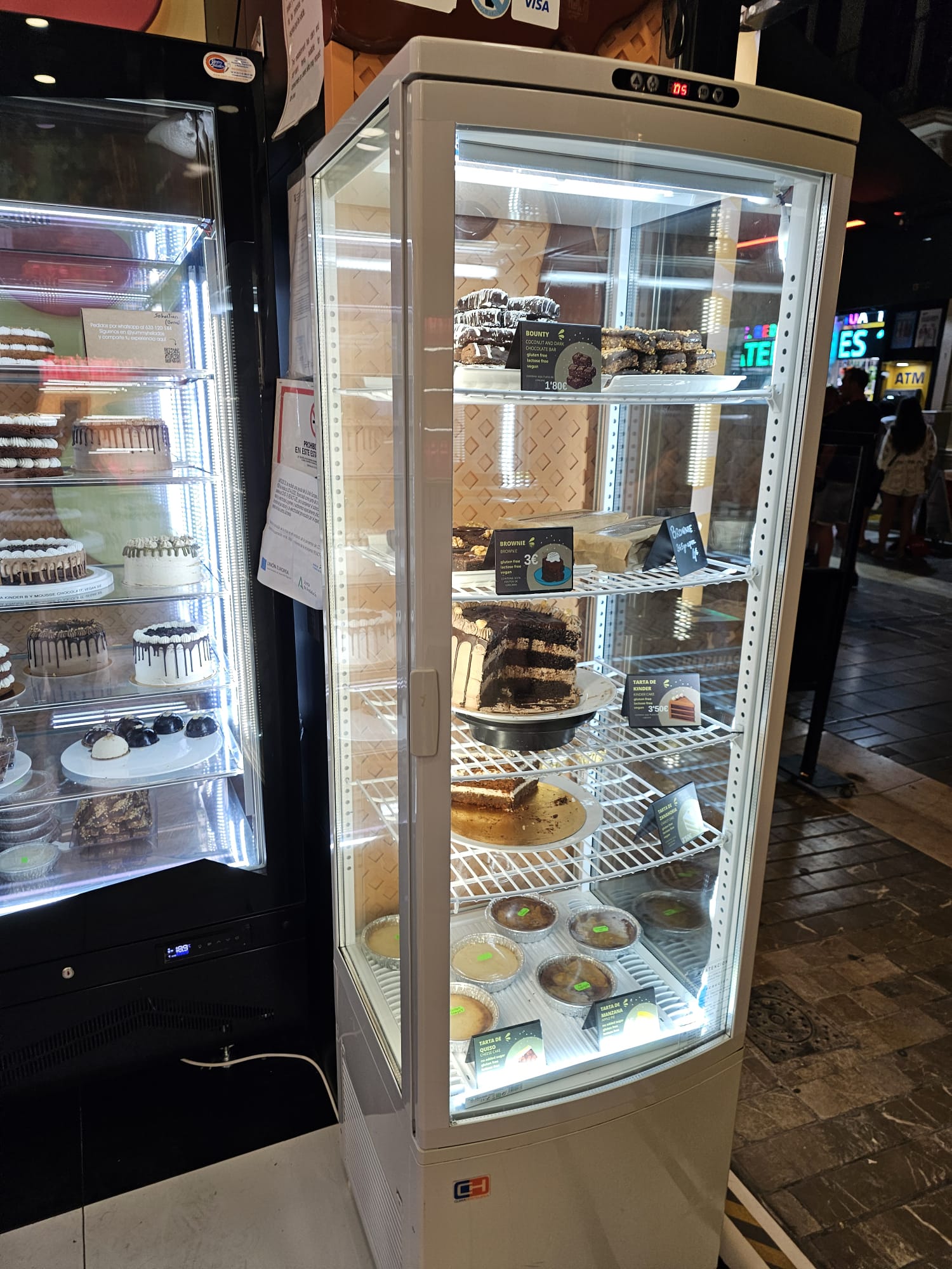 A vertical view of a bakery pastry case filled with various gluten-free treats, including pink frosted donuts, chocolate-covered pastries, and slices of cake.