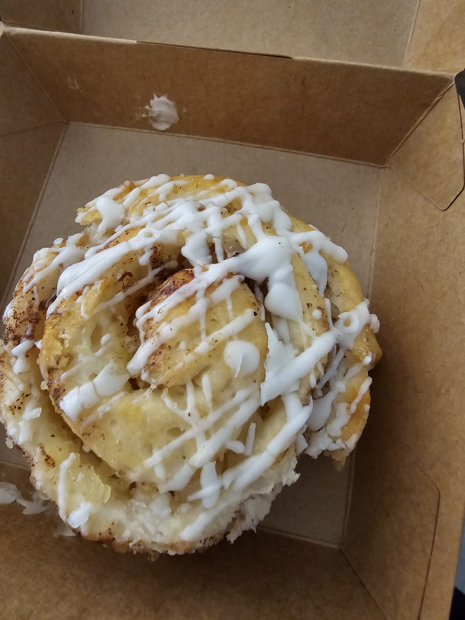 a gluten-free cinnamon roll with white icing drizzle in a cardboard box