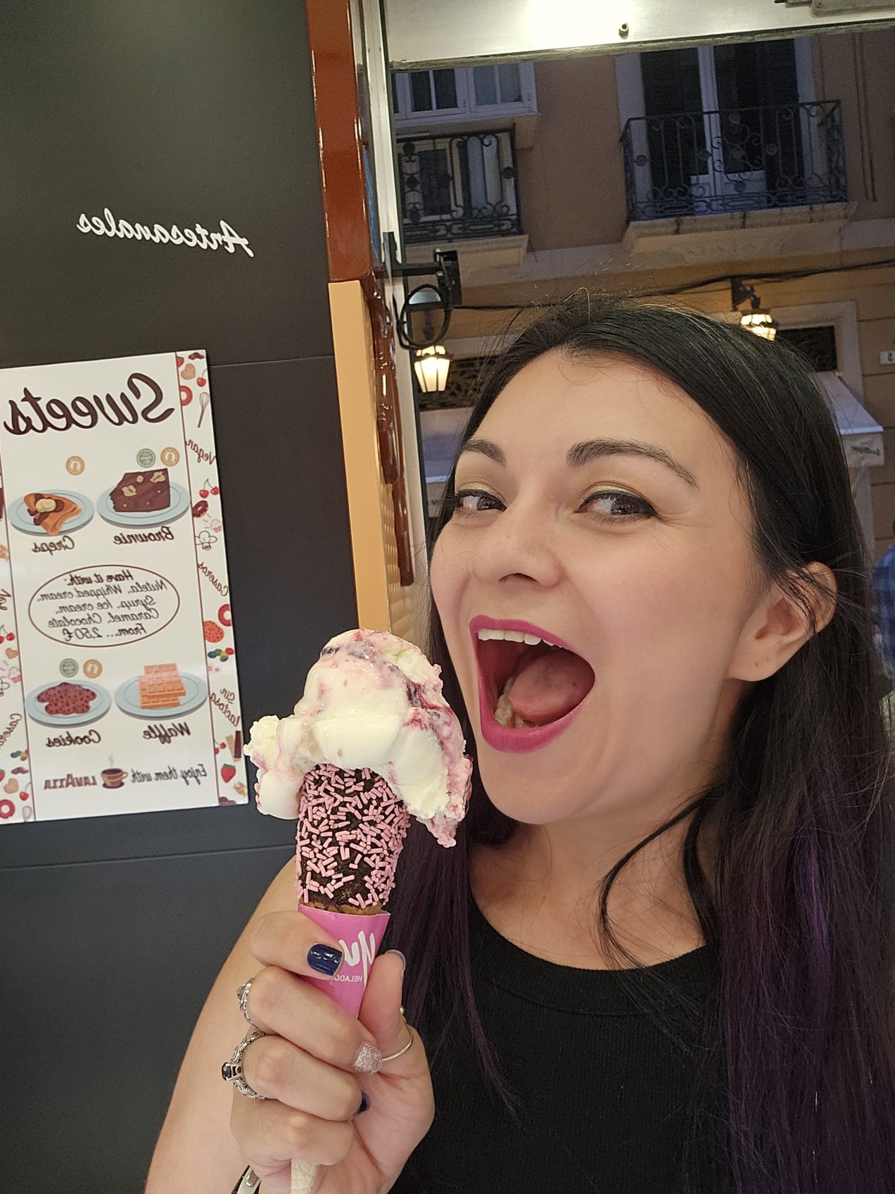 A woman happily holding a large ice cream cone with white and pink scoops, topped with chocolate coating and pink sprinkles.