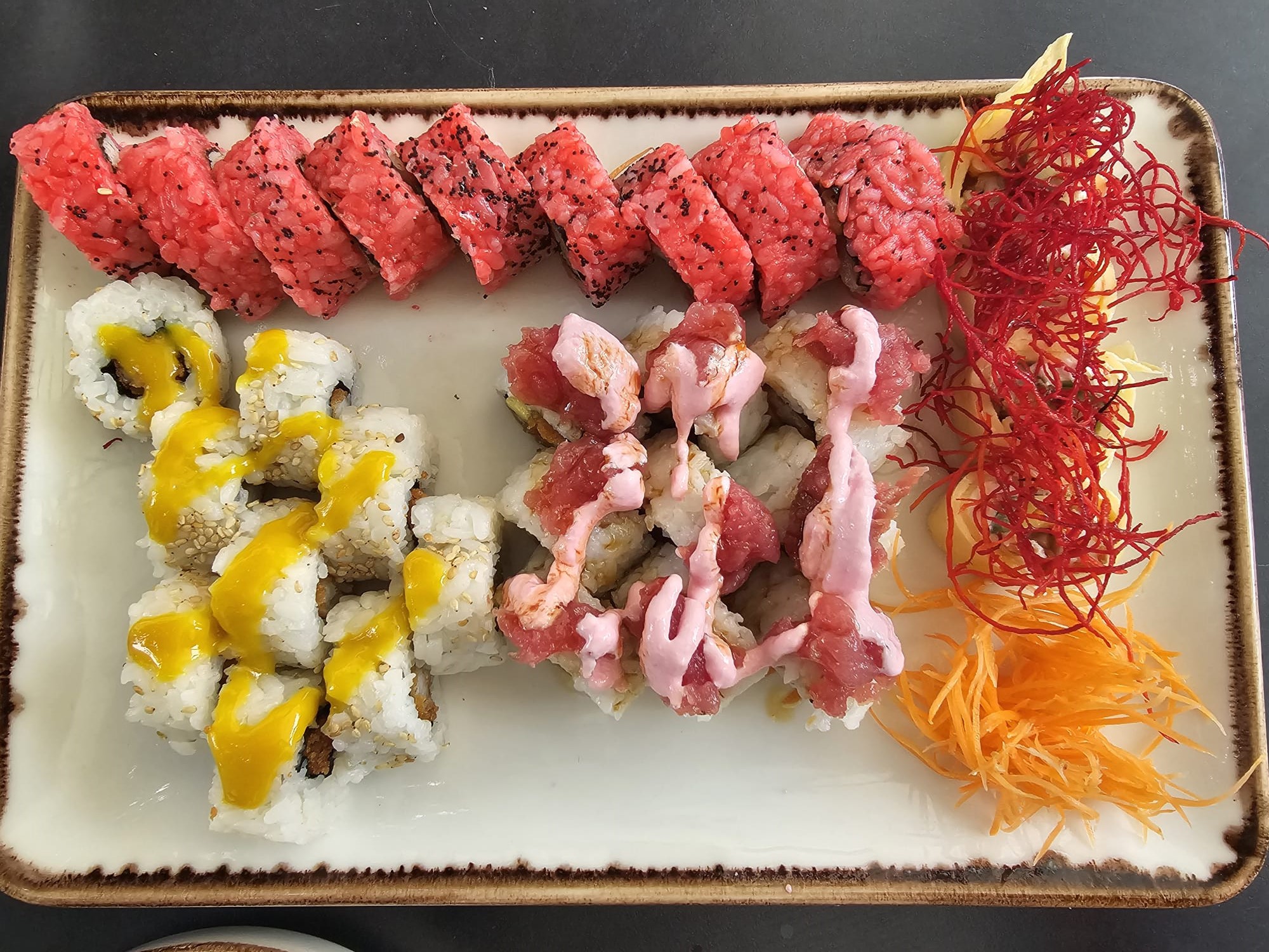 A rectangular platter of colorful sushi rolls, including some topped with bright yellow sauce, others with pink sauce, and a side of shredded carrots and beet garnish.