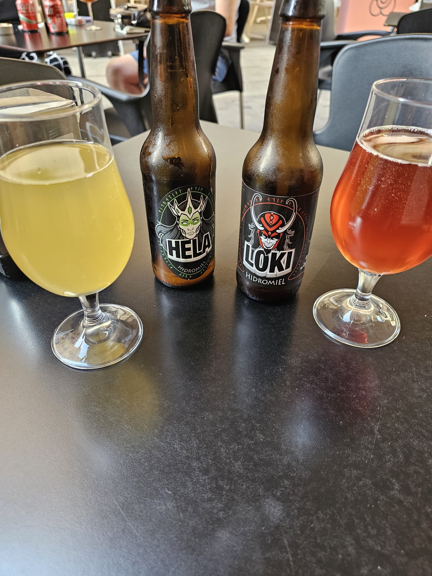 Two glasses of "Hidromel" (mead) on a table, one yellow and one amber, standing next to their respective "Hela" and "Loki" brand bottles.