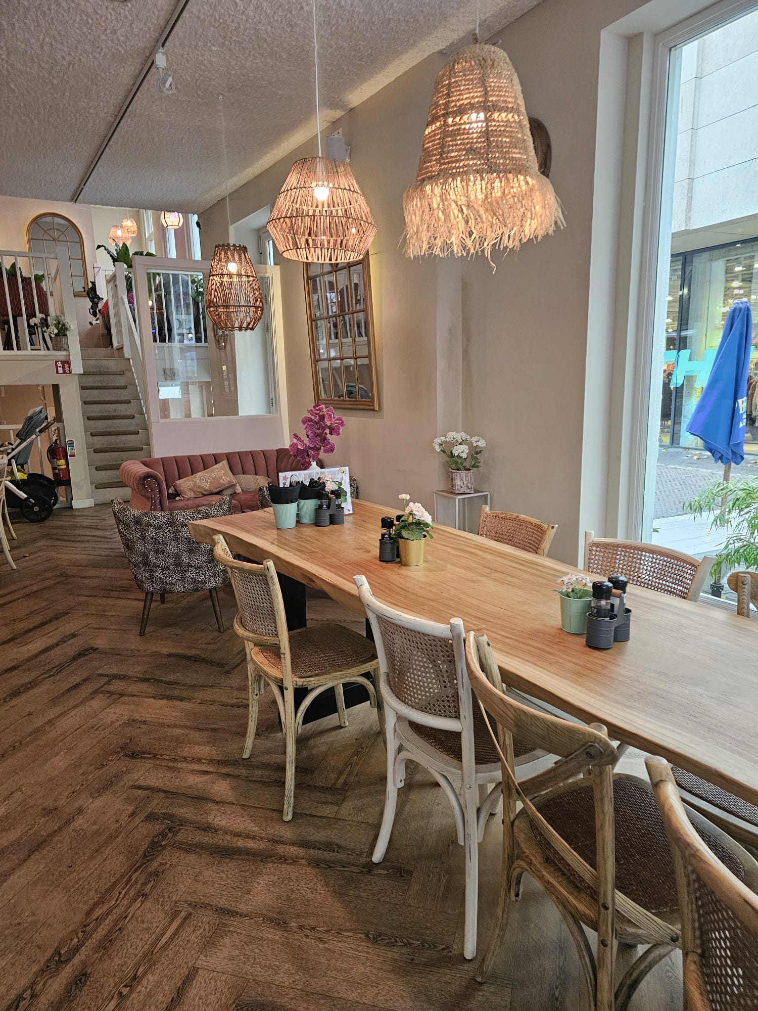 A bright, airy restaurant interior with light wood tables, white woven chairs, and bohemian-style wicker pendant lights.