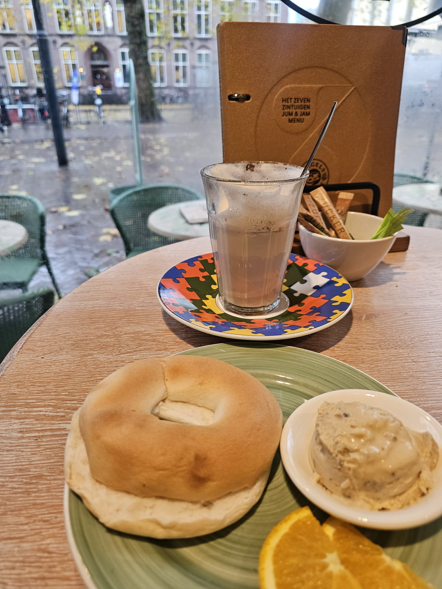 A toasted gluten-free bagel served on a green plate with a side of walnut honey cream cheese and an orange slice. A latte with colorful latte art sits in the background.